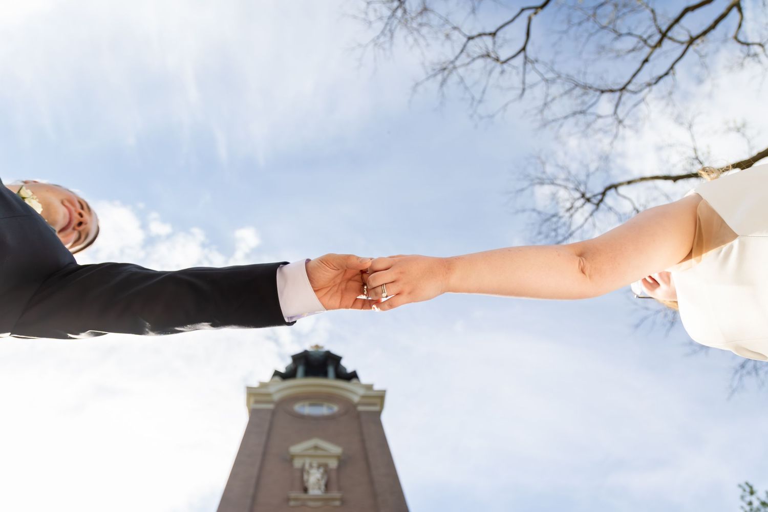 Bride and groom hold hands outdoors with historic clock tower and blue sky in background, low-angle view.