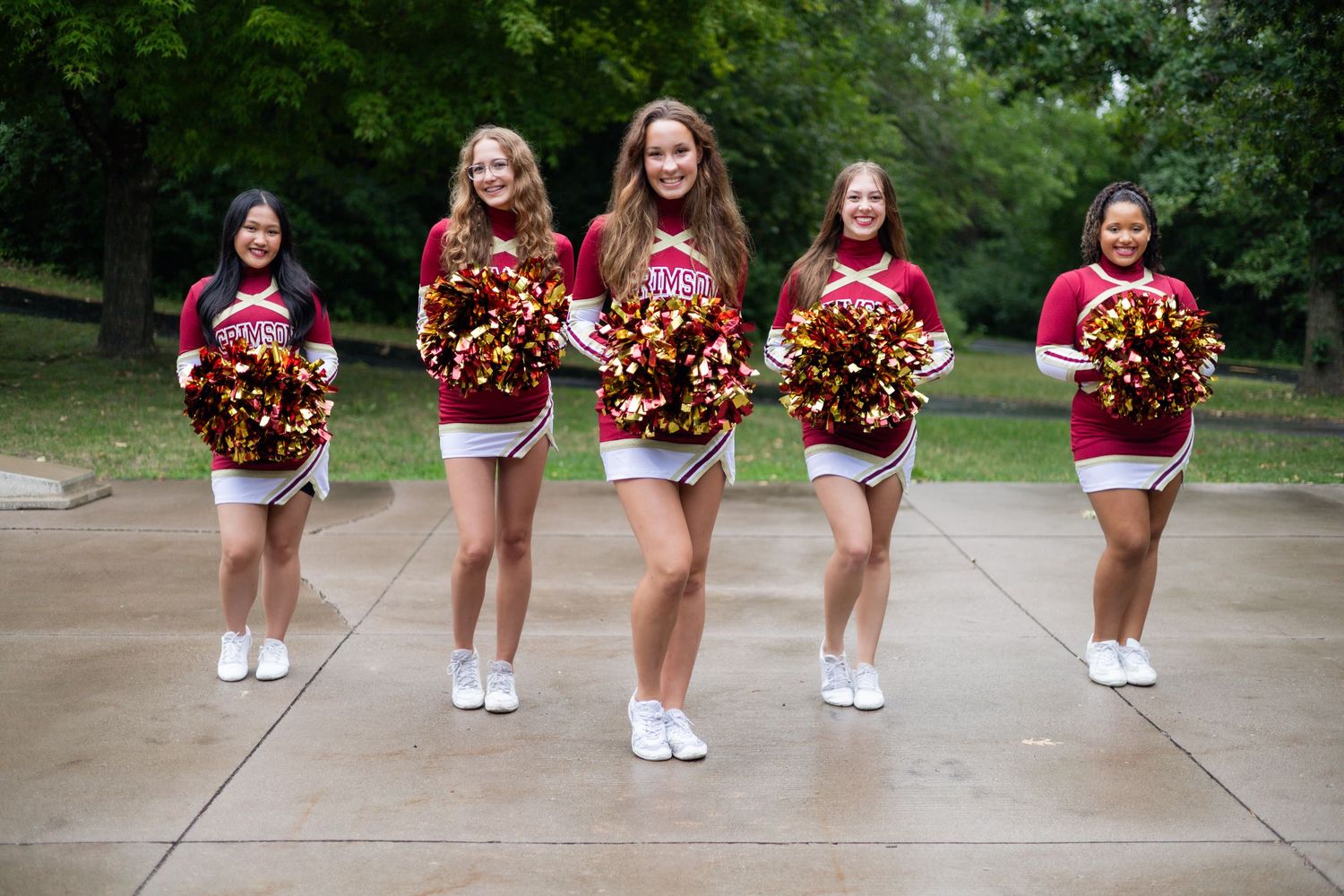 Five cheerleaders in matching red uniforms pose with pom poms on a concrete path outdoors.