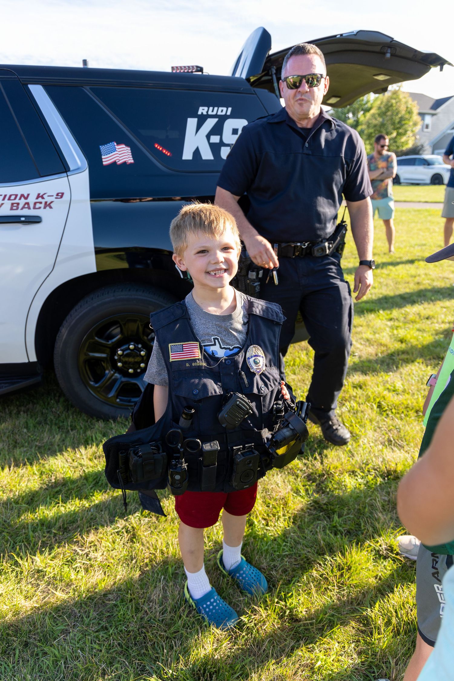 Young child holding police equipment poses next to a K-9 police vehicle on sunny grass field.