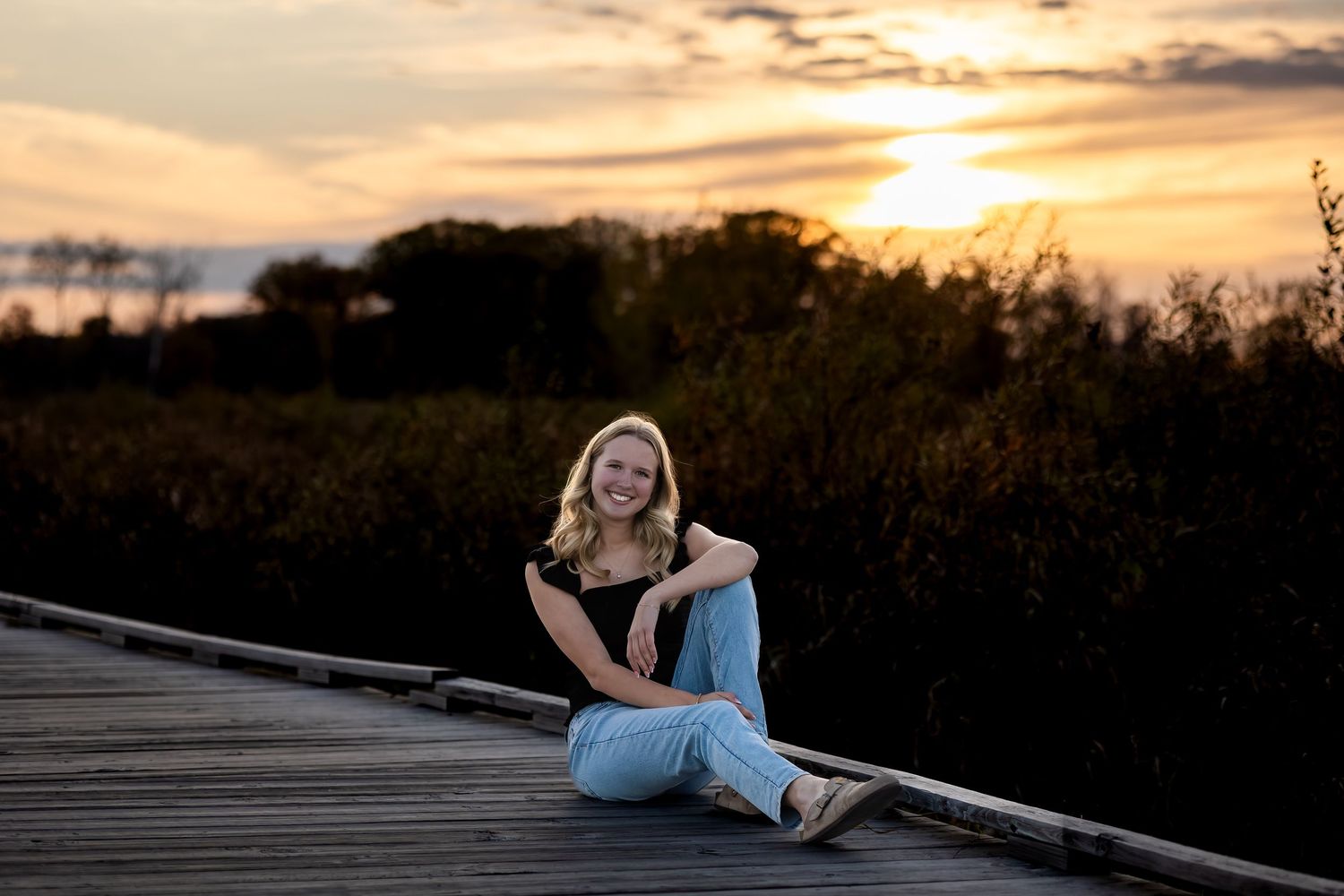 Person sitting on wooden boardwalk at sunset with dramatic golden sky and silhouetted vegetation in background.