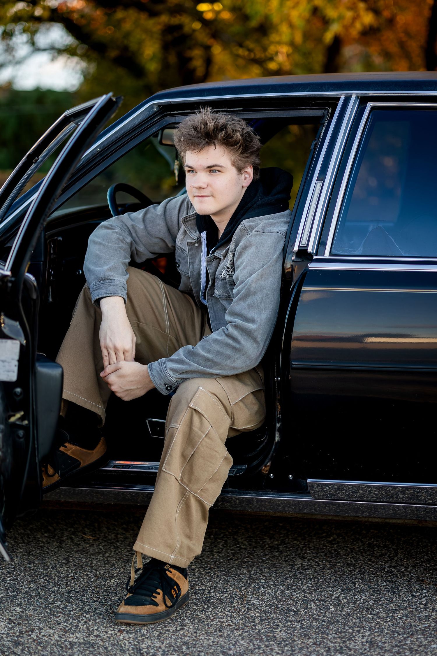 Someone sits in a black vintage car wearing a gray jacket and khaki pants during autumn.