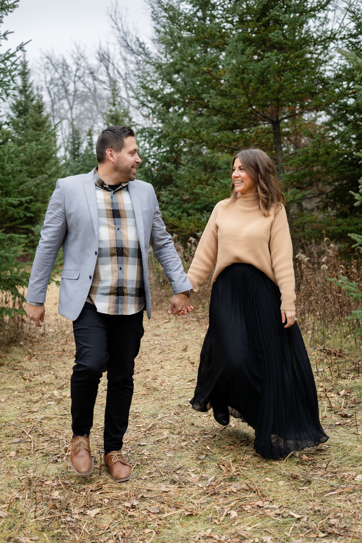 A couple in fall attire poses together in front of evergreen trees on a hillside.