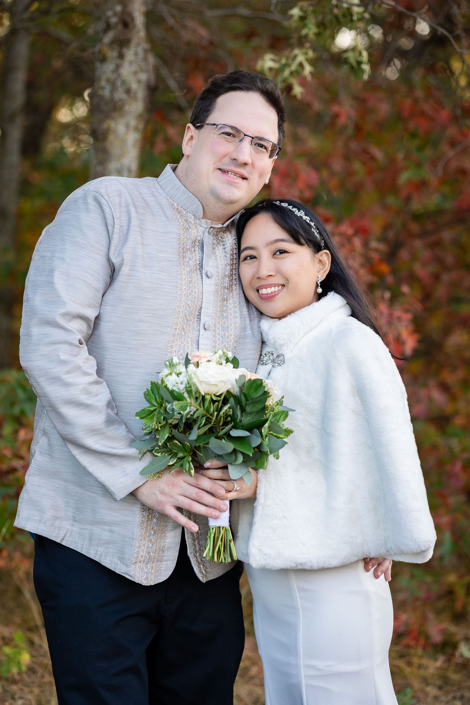 A couple poses with a white bouquet against colorful fall foliage.