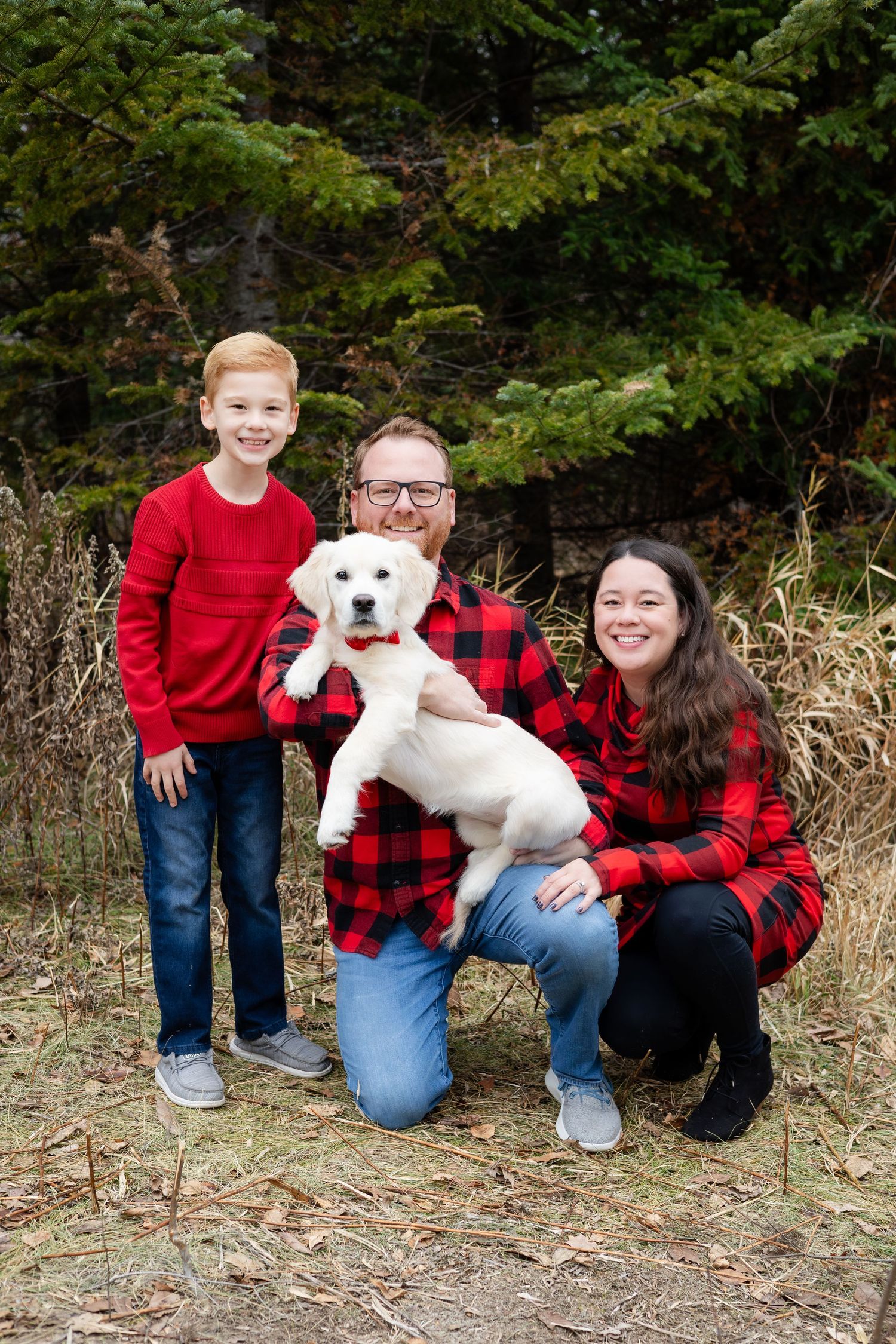 A family in coordinating red outfits sits together with their white dog in front of evergreen trees.