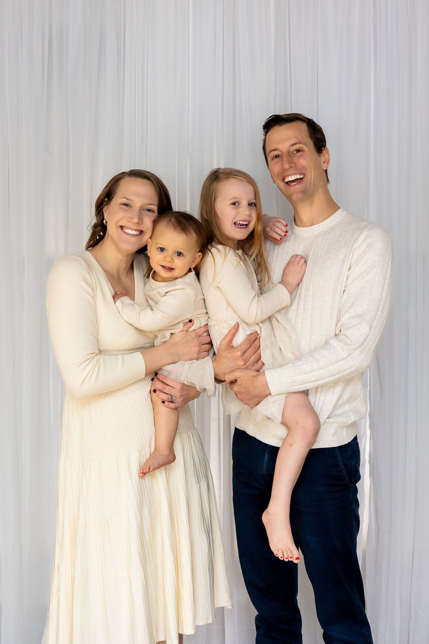 Happy family of four in matching cream outfits posing together, parents holding two young children, smiling against white backdrop.