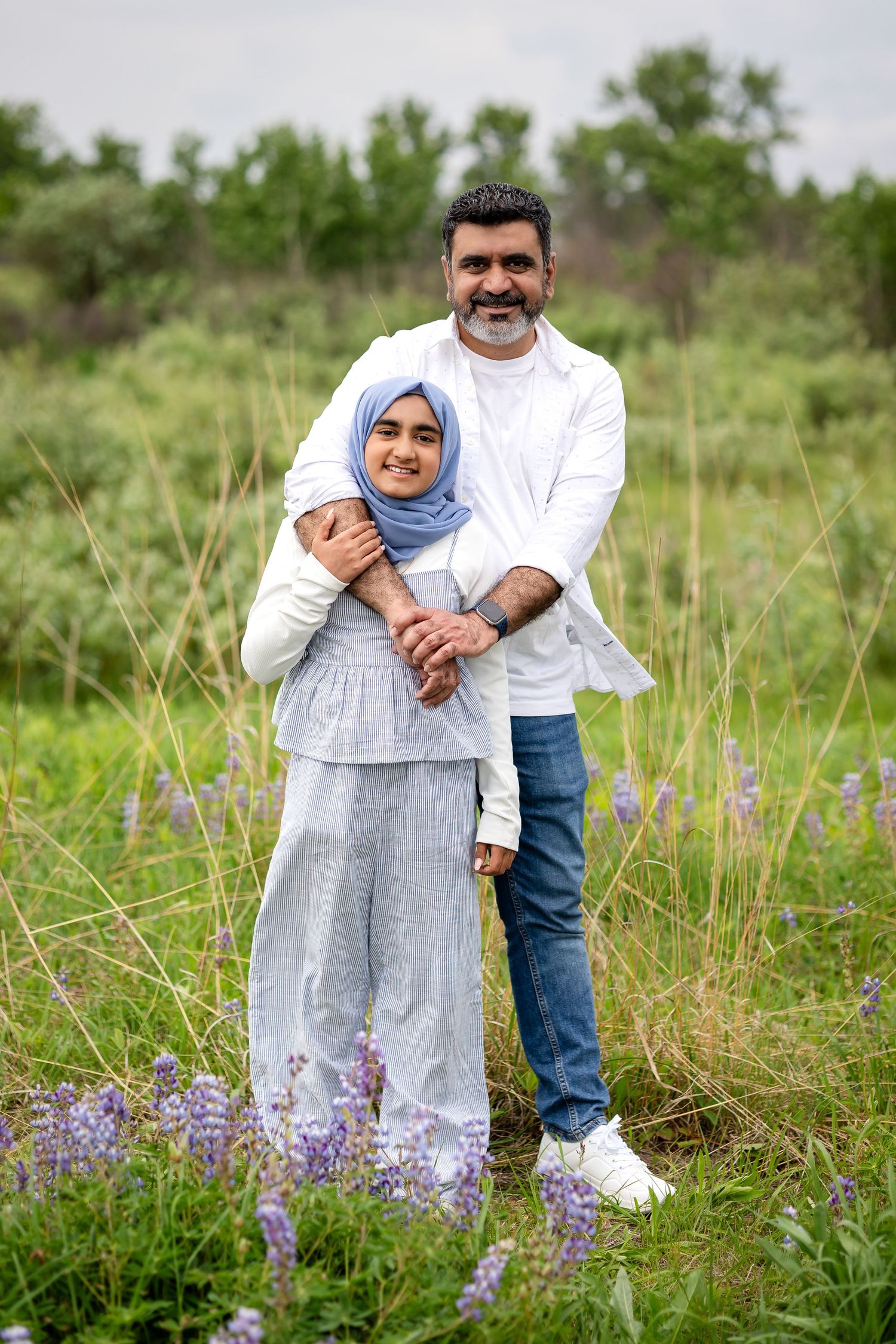 A couple stands embracing in a green field with purple wildflowers, the woman wearing a blue hijab and gray outfit.