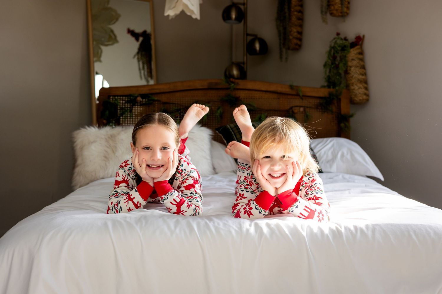 Two children in matching Christmas pajamas lying on a white bed, smiling and posing for a holiday photo.