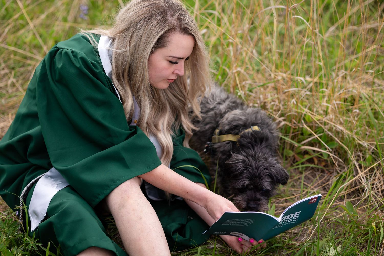 Blonde graduate in green gown sitting in grass reading diploma with her small gray dog.