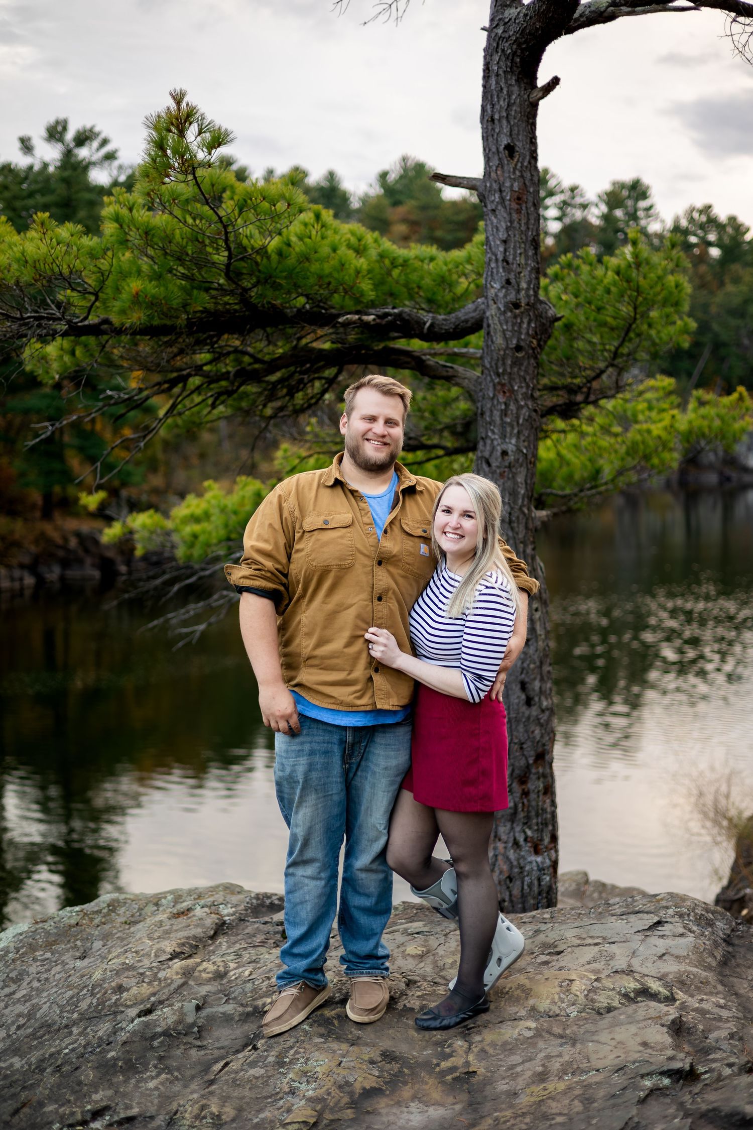 Smiling couple standing by lakeside tree in autumn, woman in striped top and red skirt.