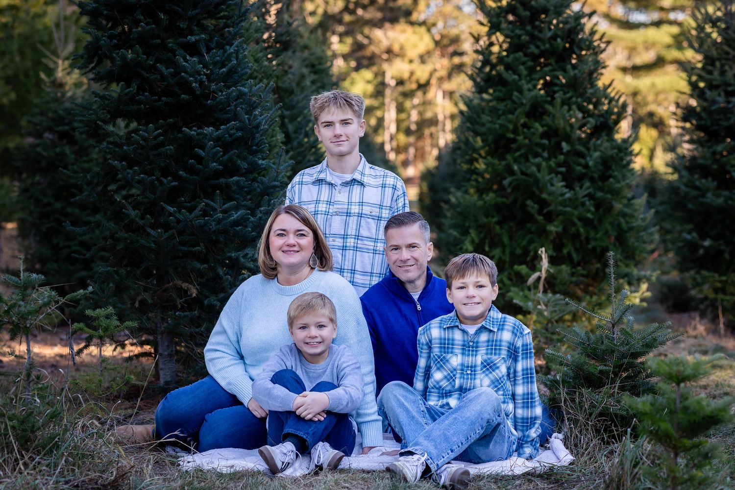 Family wearing coordinated blue and white outfits poses for outdoor portraits in a wooded setting during sunset.