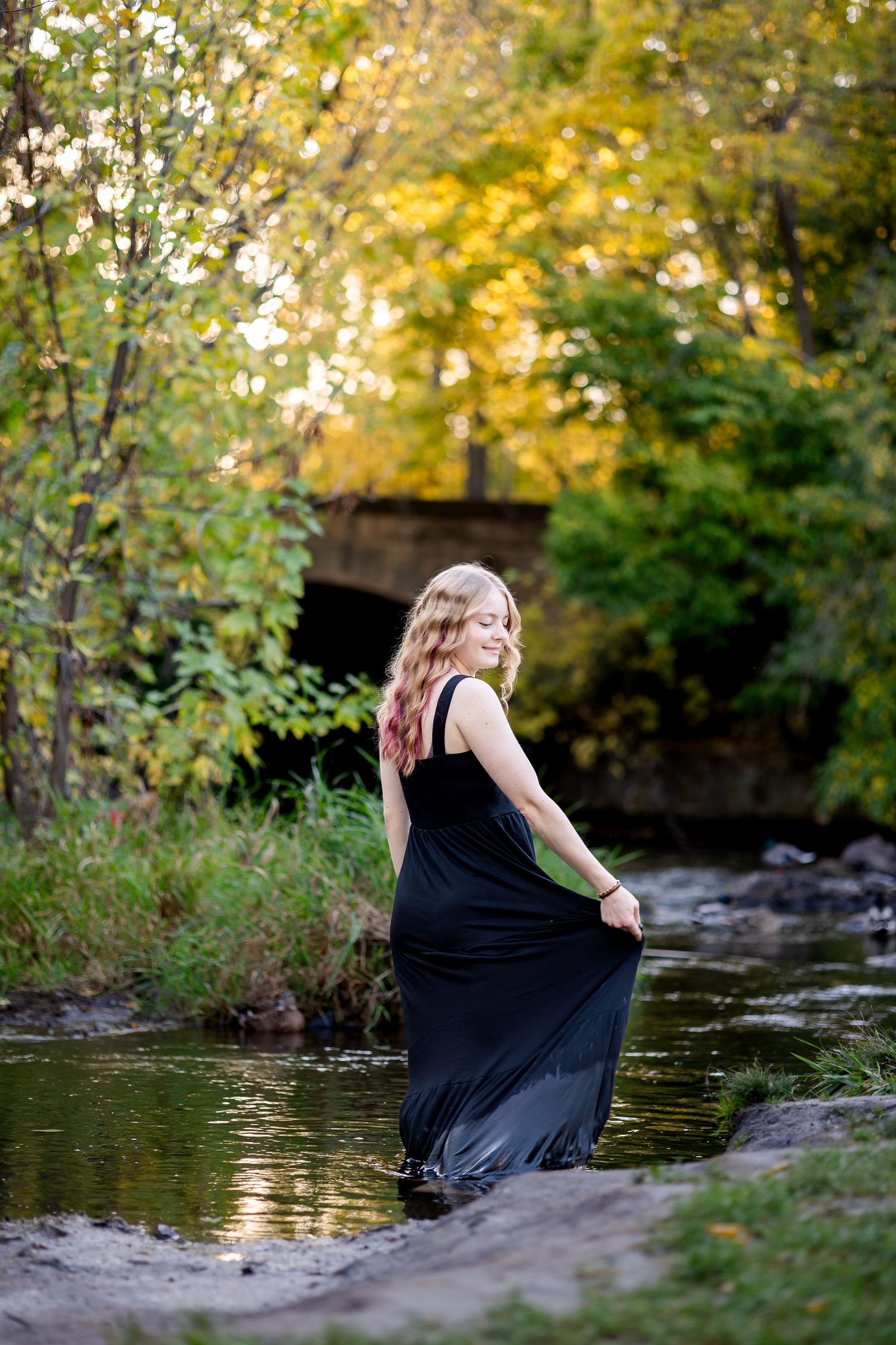 A person in a black dress sits by a tranquil stream surrounded by autumn foliage and golden sunlight.