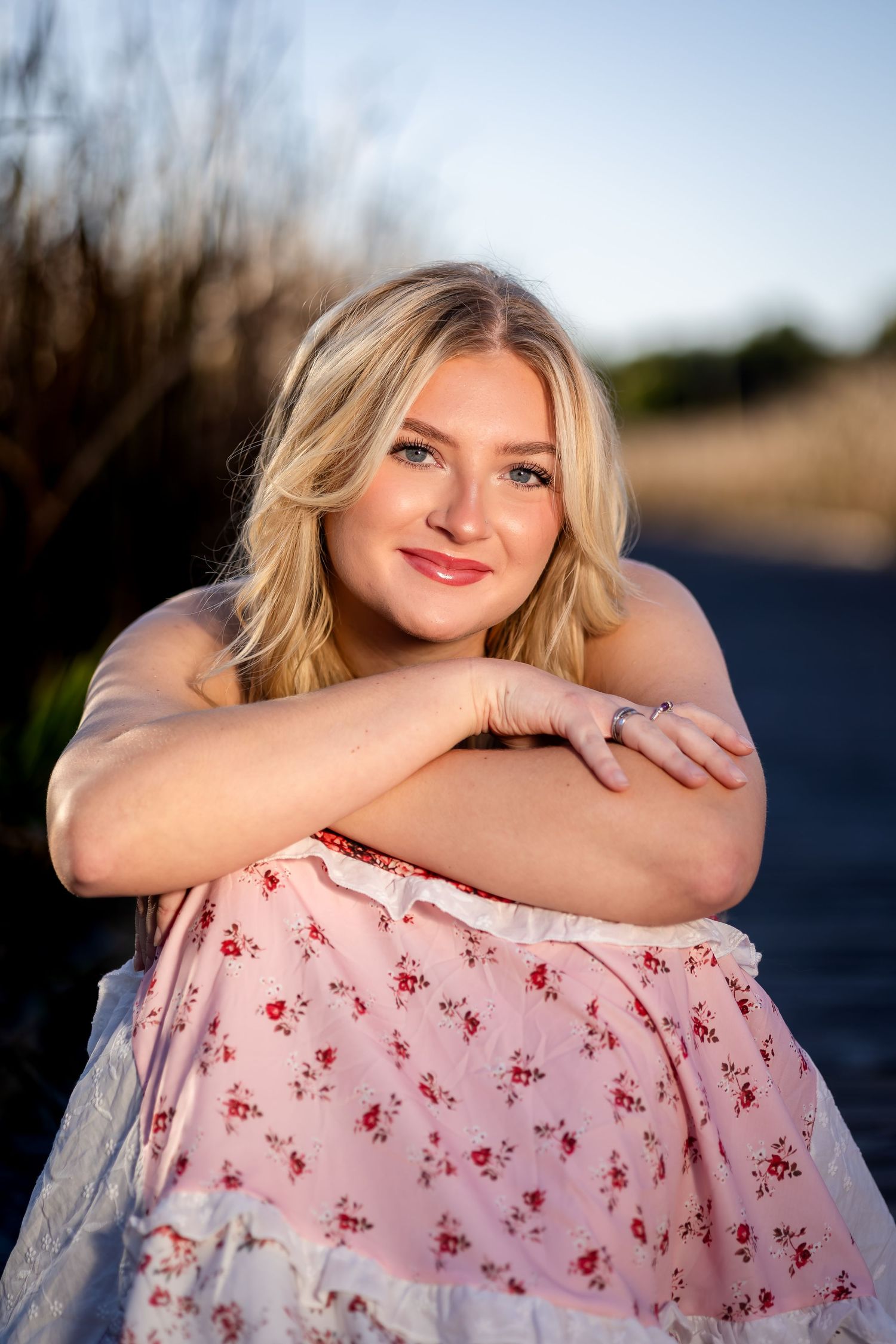 A portrait of someone in a pink floral dress sitting outdoors during golden hour with a warm smile.