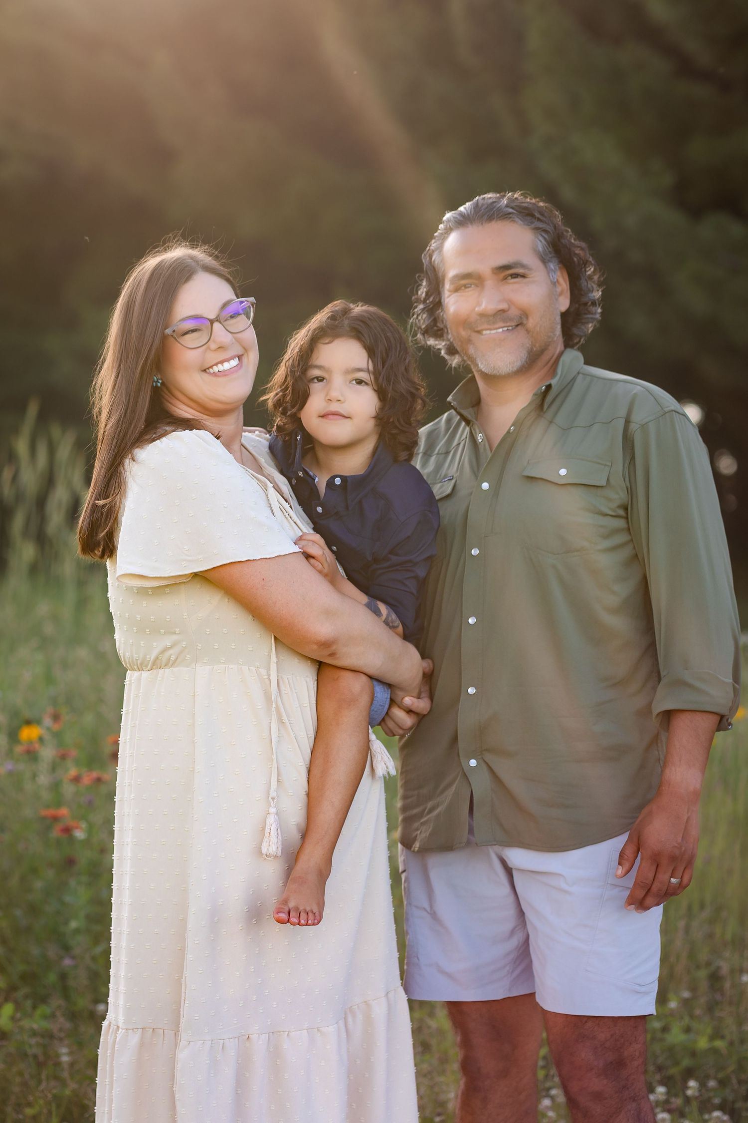 A family of three poses together outdoors in a meadow at sunset wearing casual clothing.