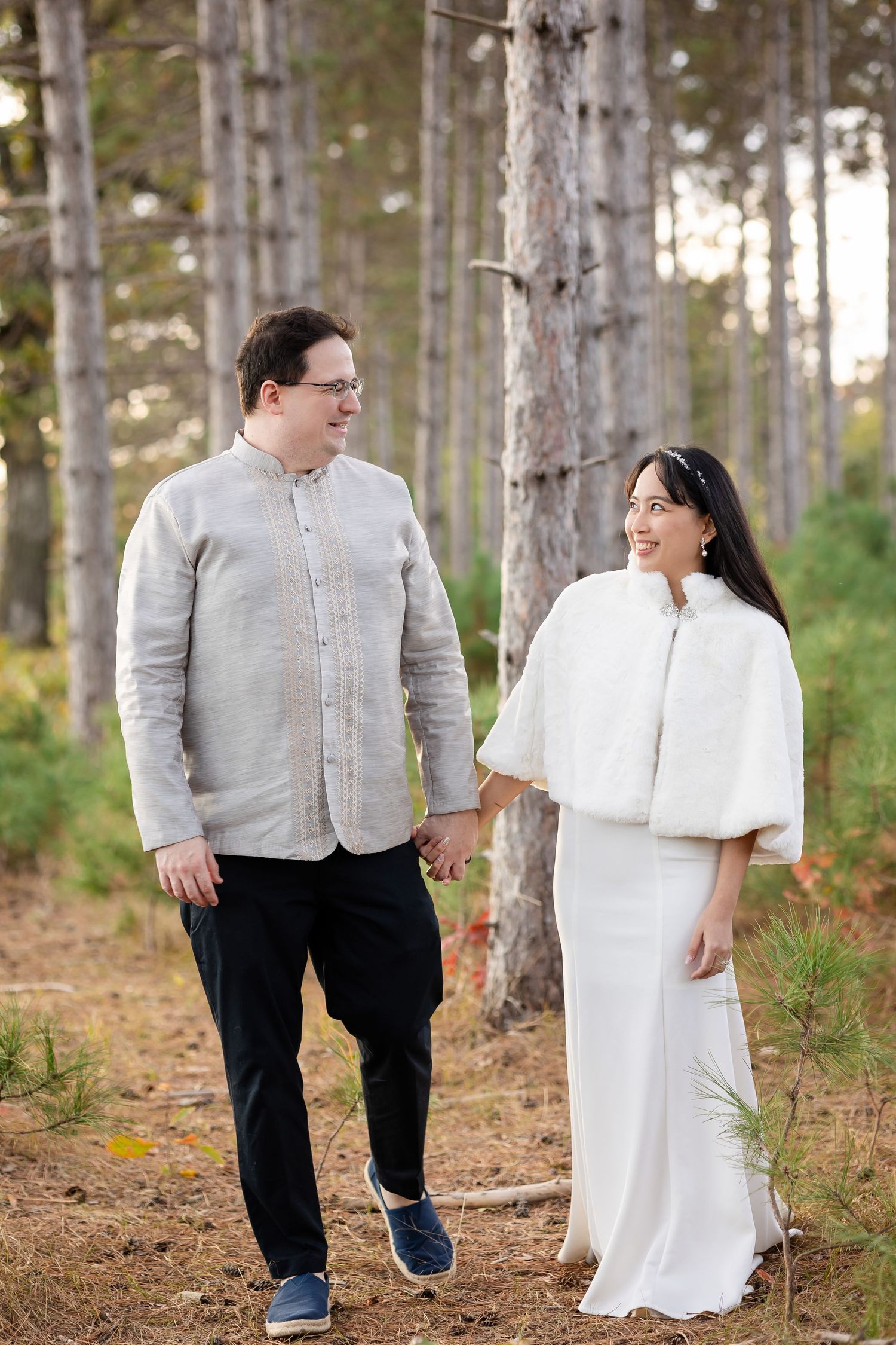 A couple walking hand in hand through a pine forest in wedding attire.