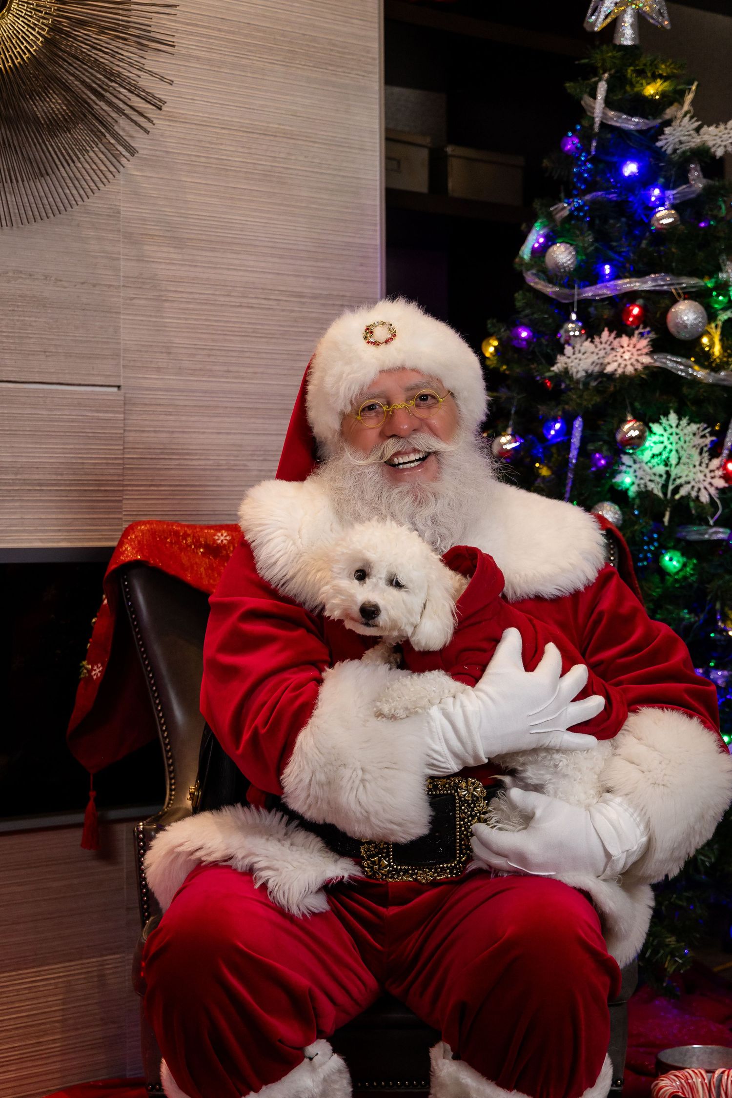 Santa Claus laughs heartily while seated in his chair next to a beautifully lit Christmas tree.