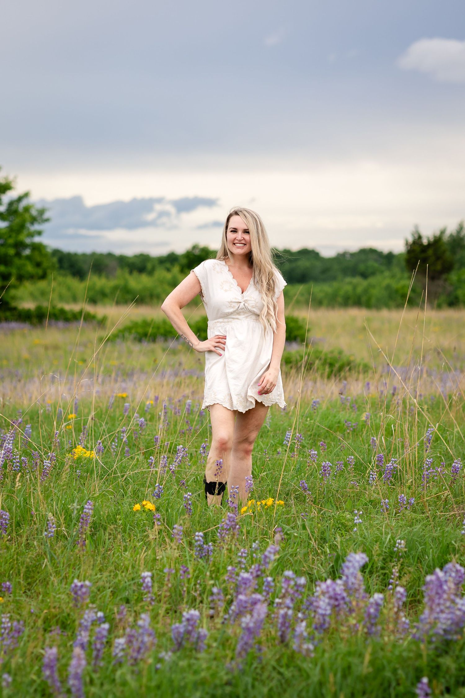 Smiling blonde woman in white dress and boots standing in a wildflower meadow with purple lupines under cloudy sky.