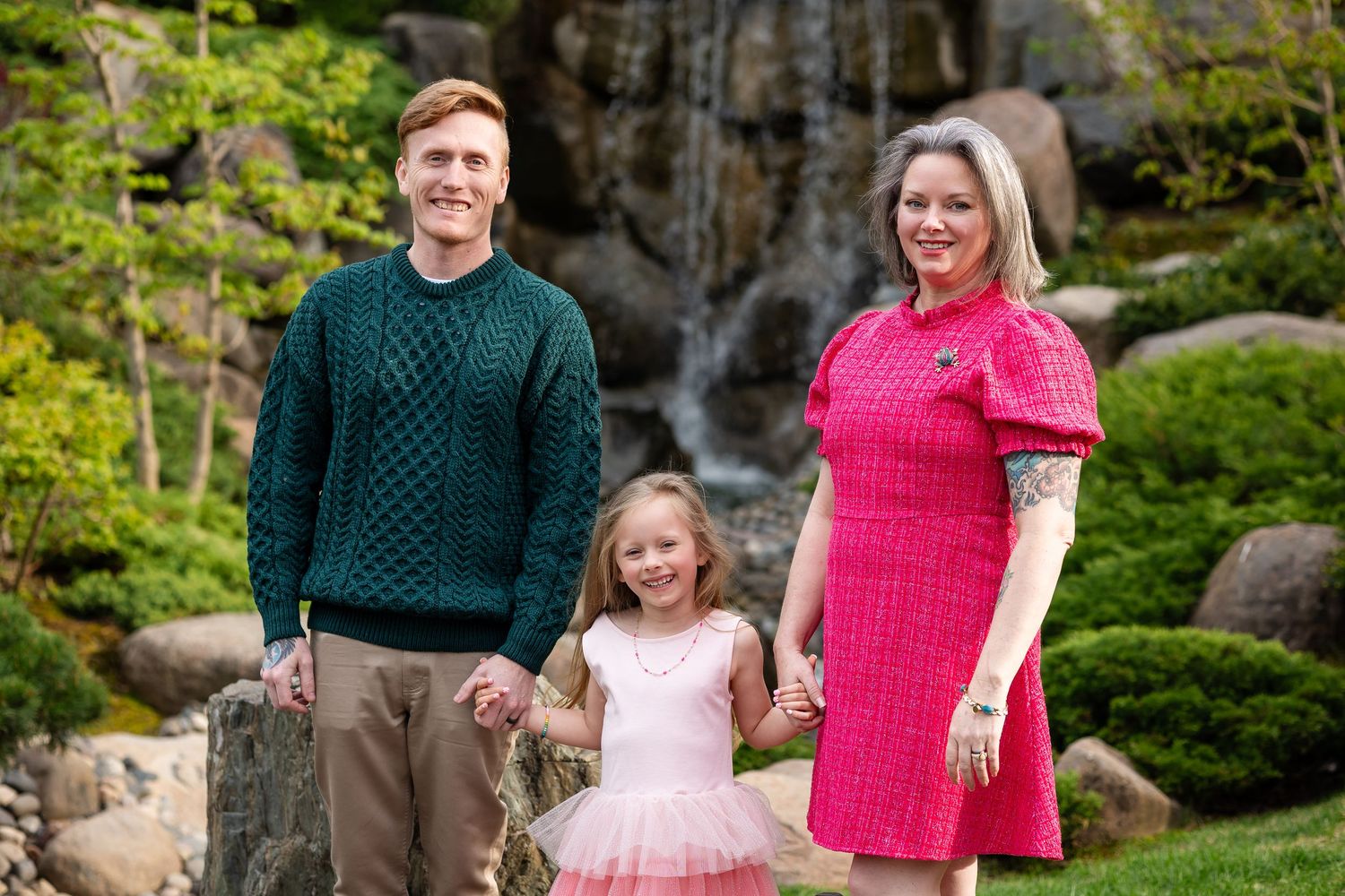 Family of three posing outdoors near a waterfall, parents holding daughter's hands in a lush garden setting.