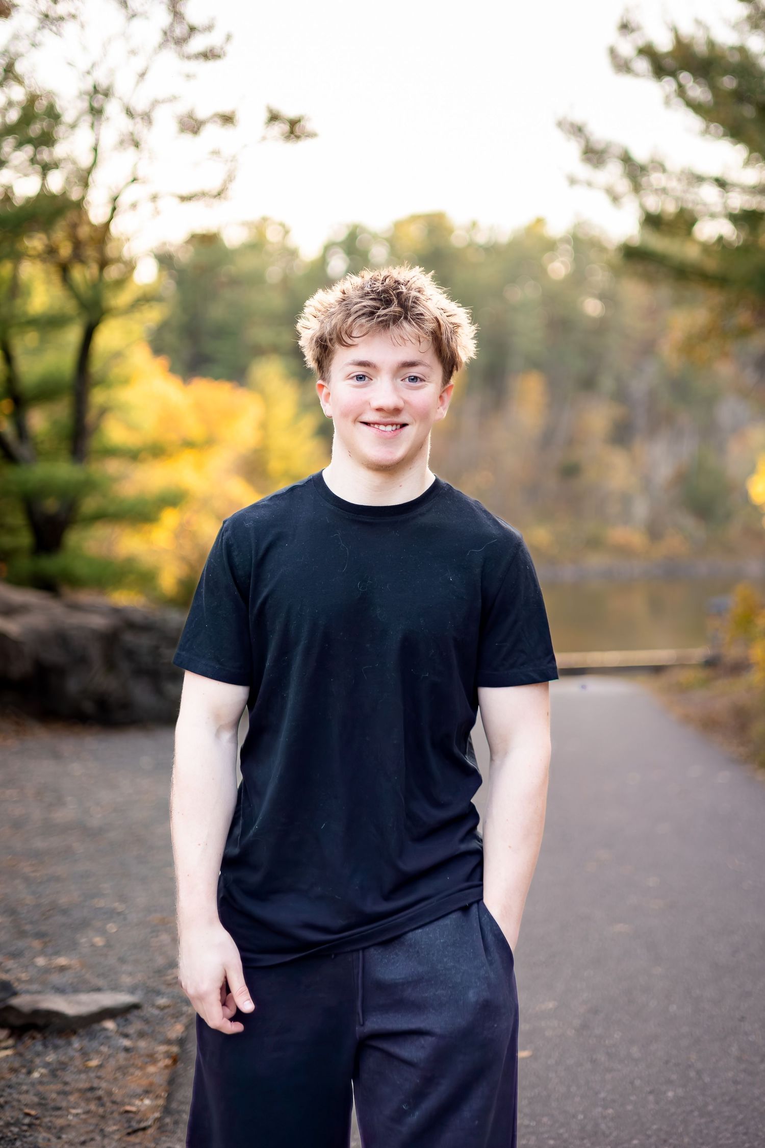 A person wearing a black t-shirt stands on a paved path surrounded by autumn trees and fallen leaves.