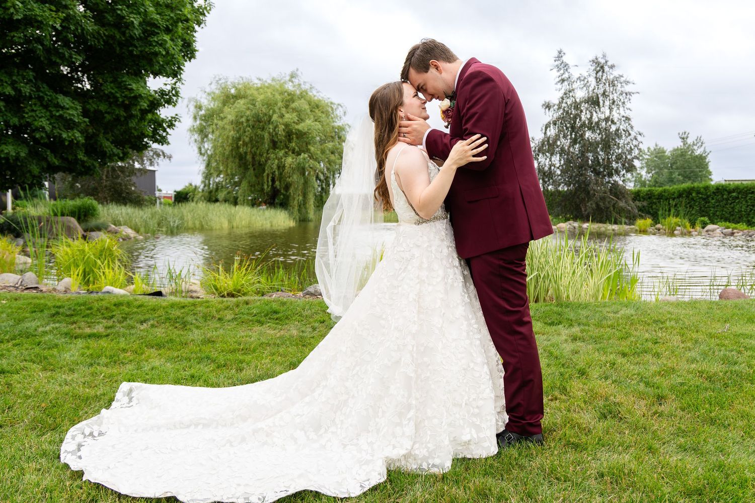 Wedding couple poses on green lawn near water with elegant white dress and burgundy suit.