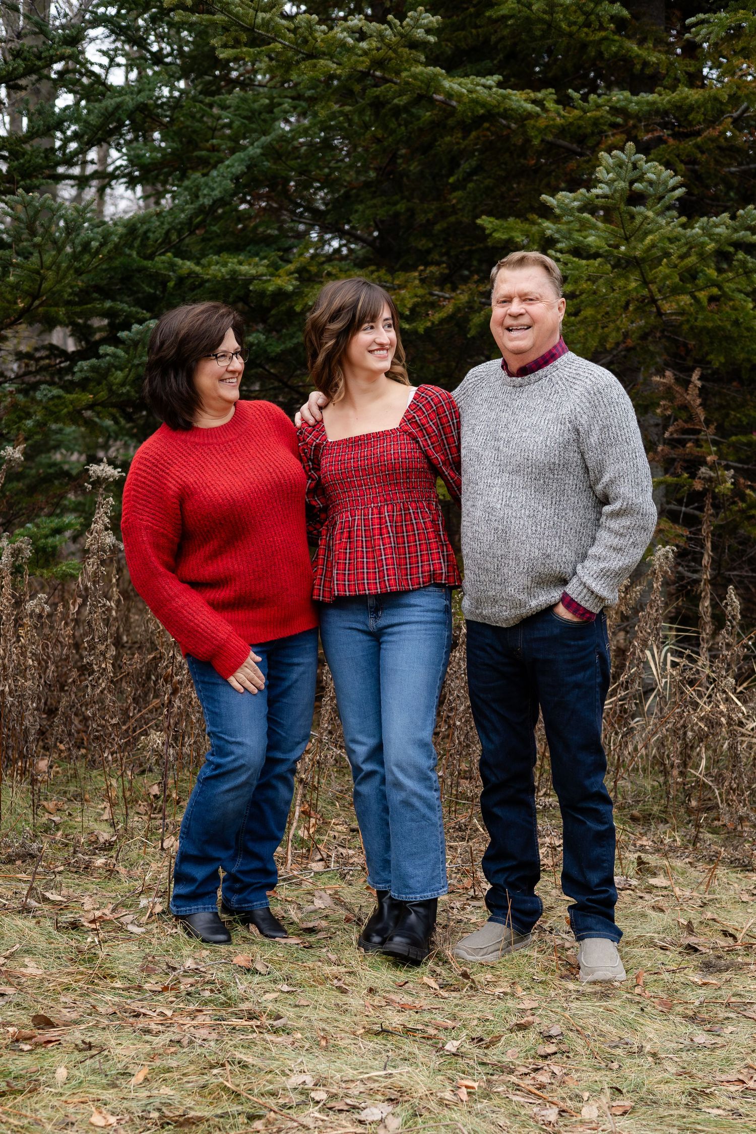 Three people in coordinated red and gray outfits stand together against a backdrop of evergreen trees.