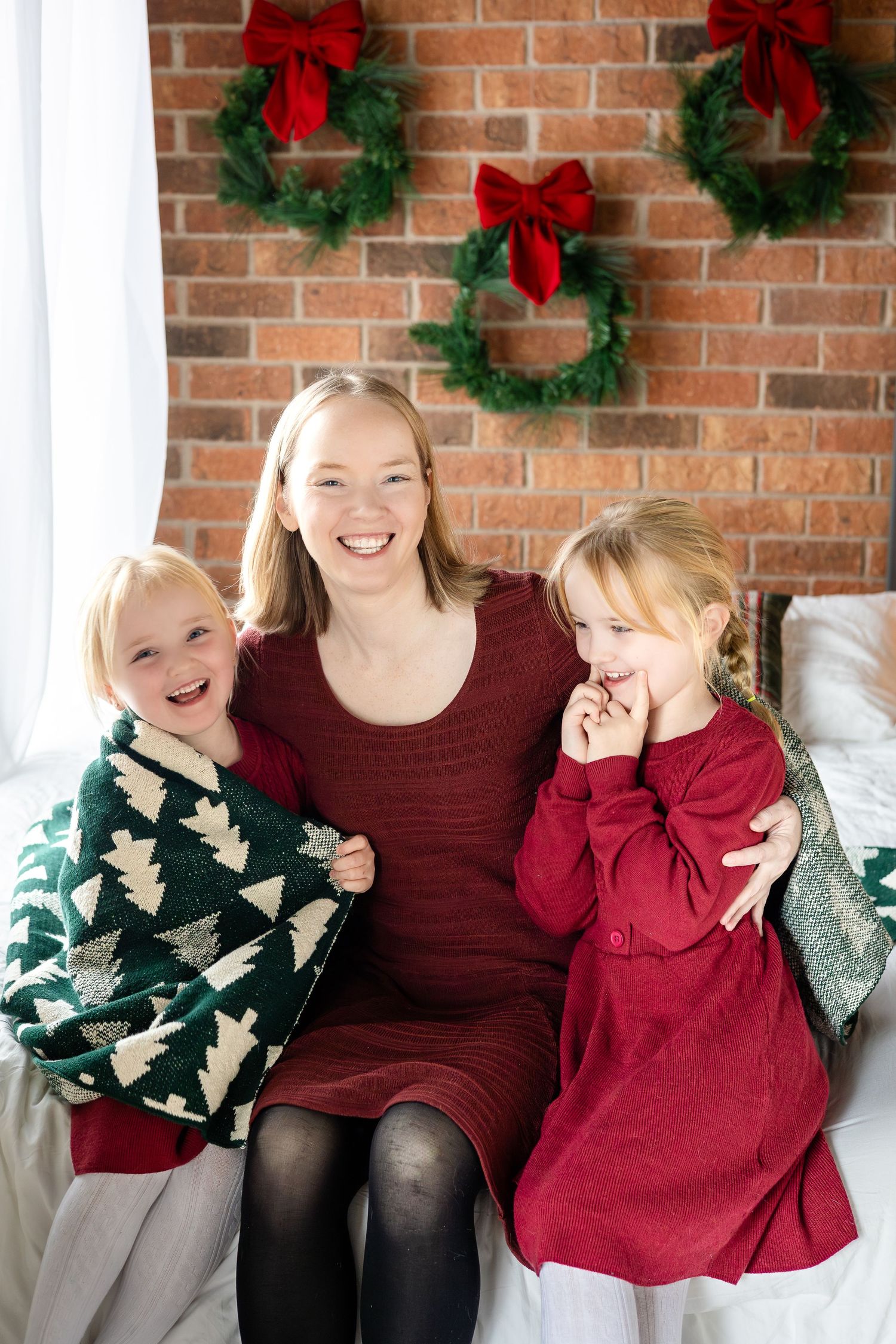 Family wearing burgundy and green sweaters pose in front of brick wall with Christmas wreaths.