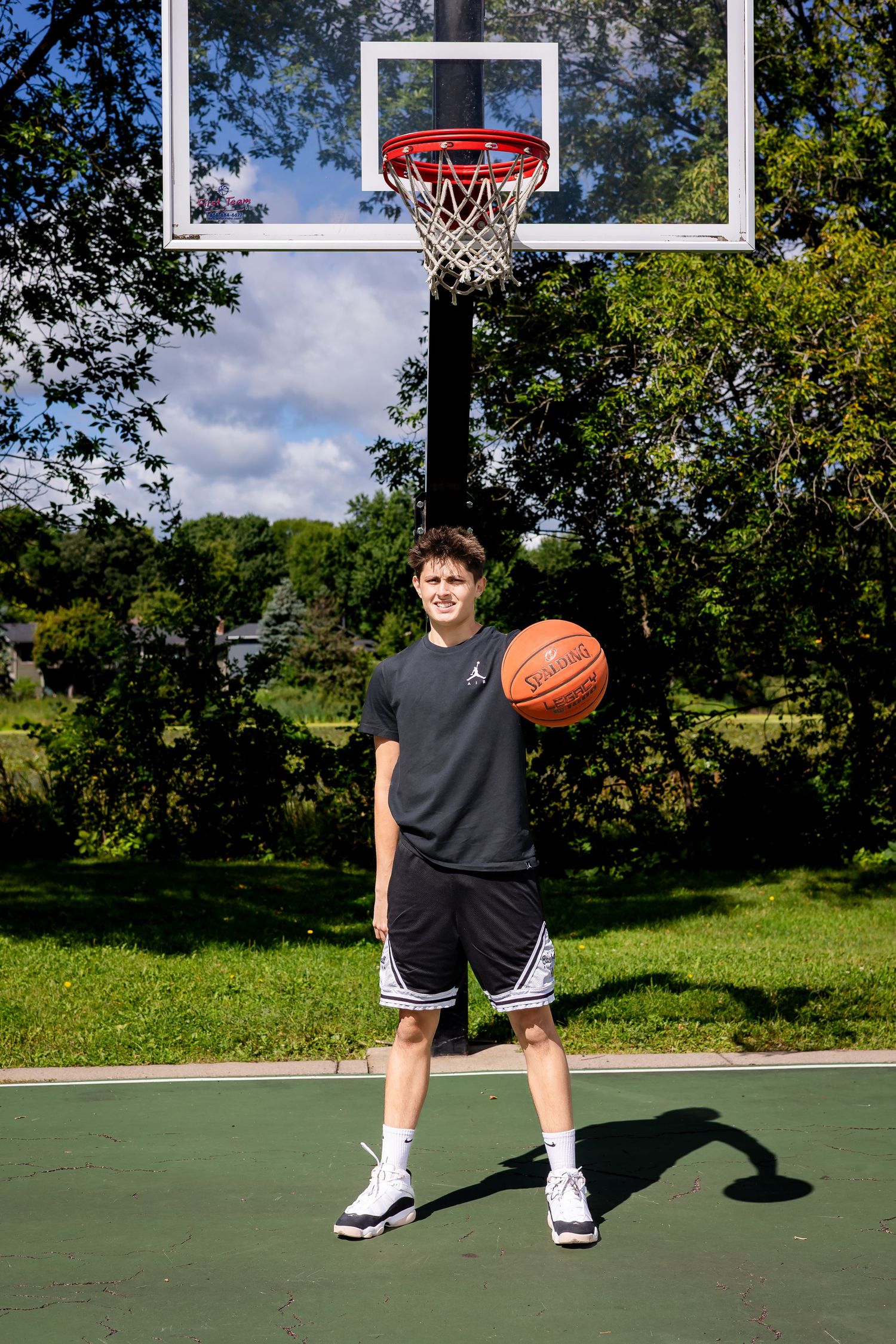 Player demonstrating basketball stance on green outdoor court with trees in background.
