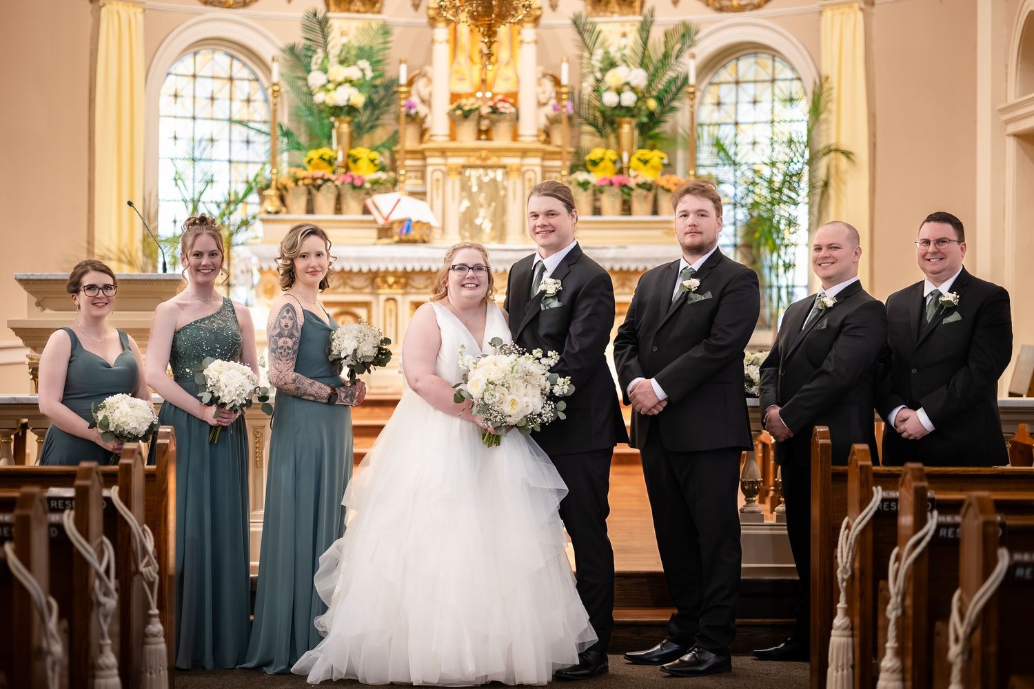 Wedding party poses in elegant church with bride in white gown, bridesmaids in teal dresses, and groomsmen in black suits.