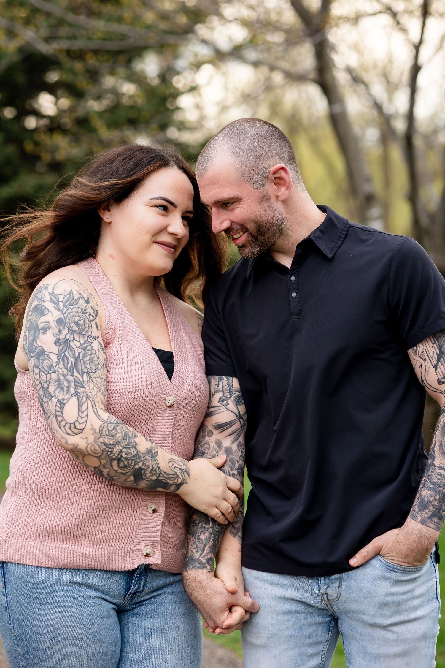 A tattooed couple holds hands and smiles at each other outdoors, dressed casually in jeans and a pink cardigan.