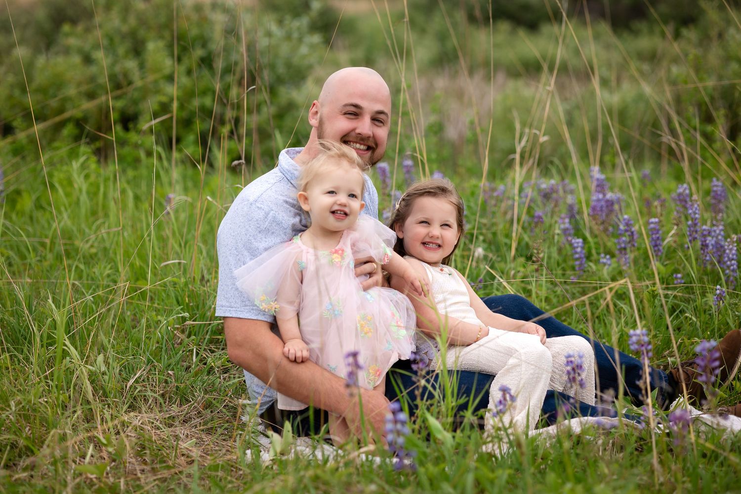 Bald father smiling with two young daughters in a wildflower meadow with purple lupines and tall green grass.