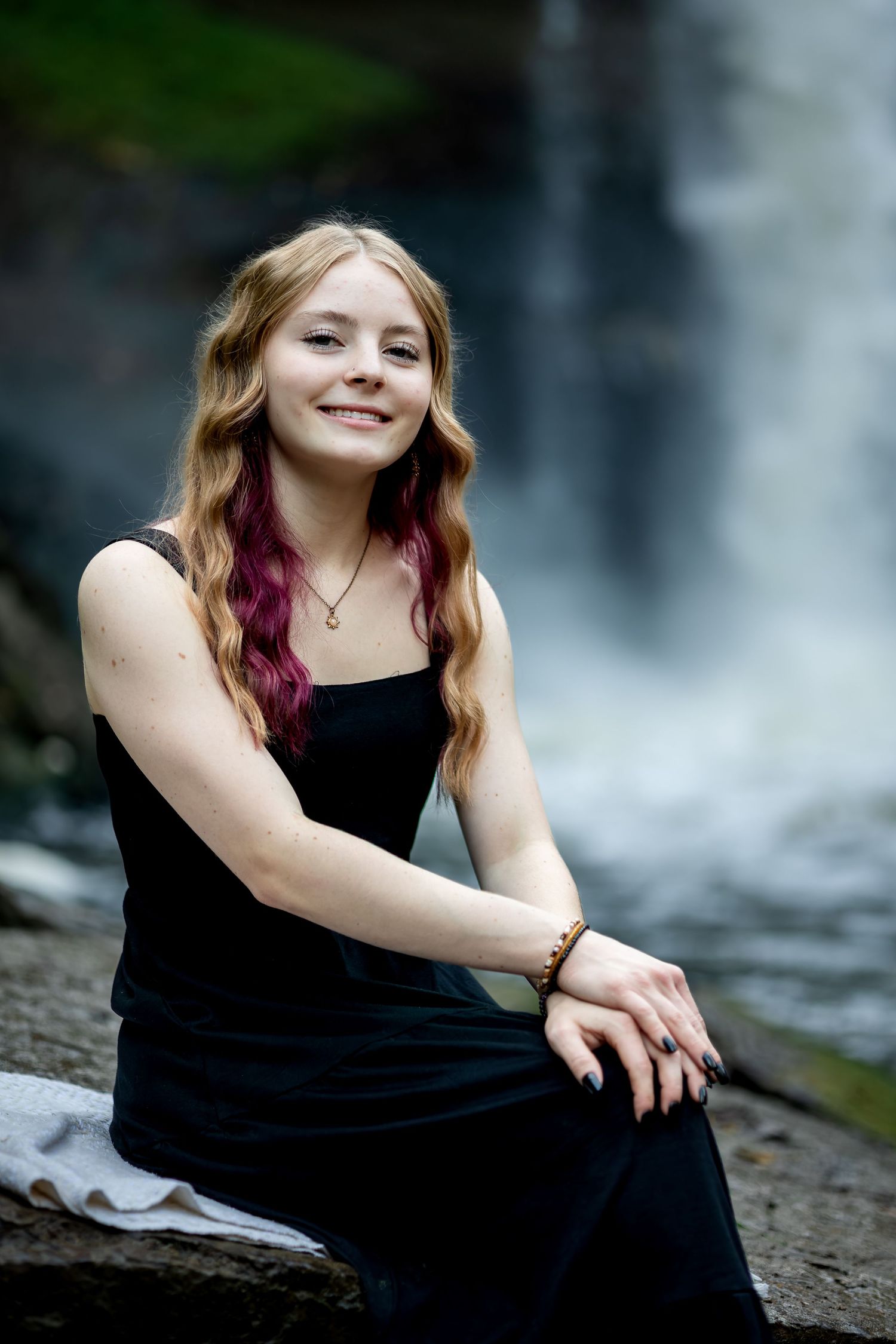 A figure in a black dress poses gracefully near a cascading waterfall in a natural outdoor setting.