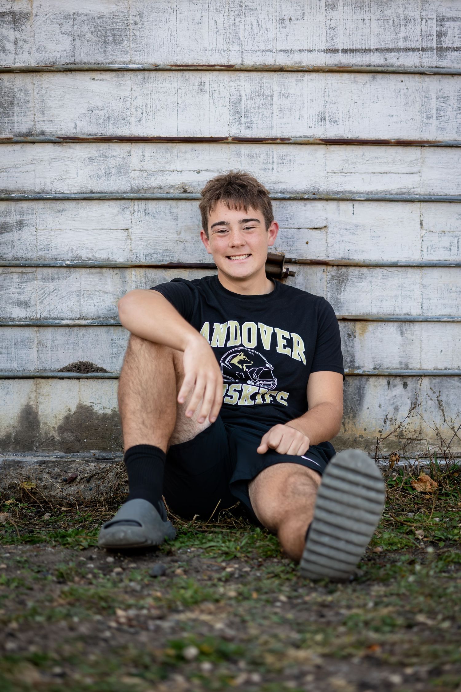 A person in a black t-shirt sits against a weathered white wooden wall outdoors.