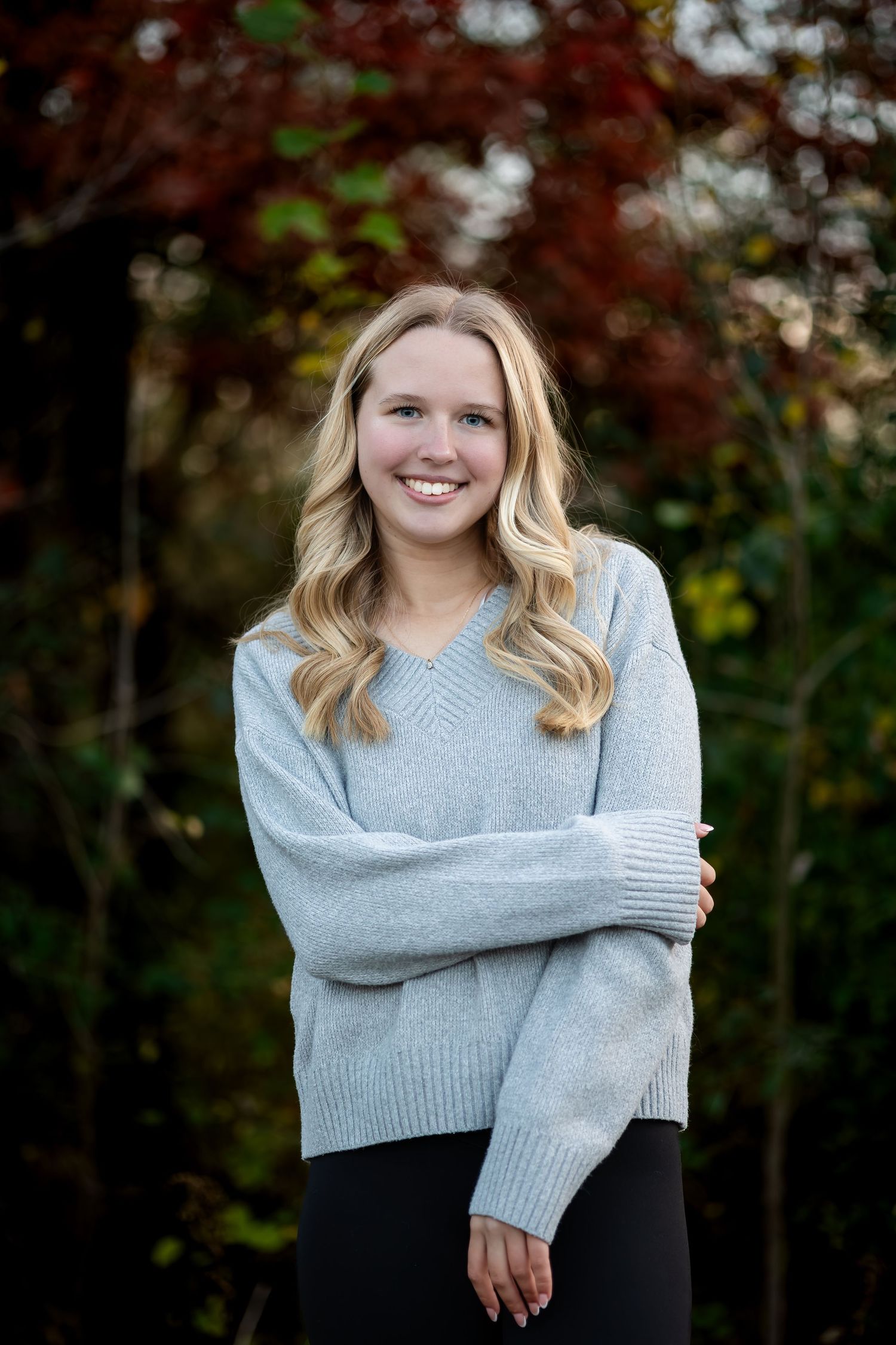A person wearing a gray sweater poses outdoors during a fall photoshoot with natural foliage in the background.