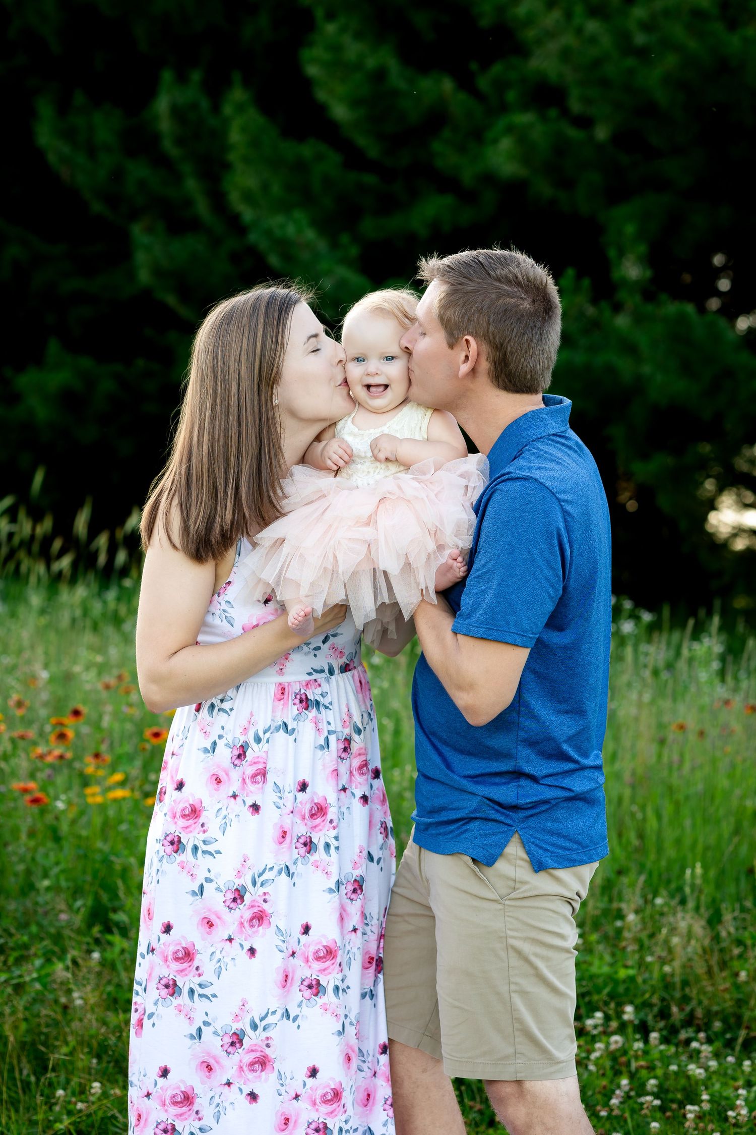 A couple shares tender moments with their baby in a lush green field with wildflowers on a sunny day.
