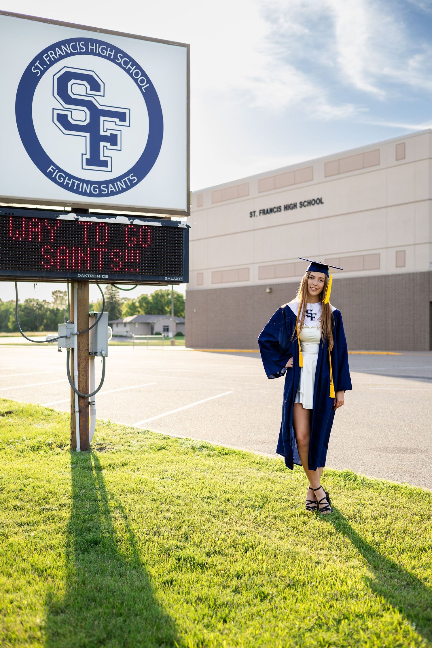Graduate in blue cap and gown poses by St. Francis High School sign.