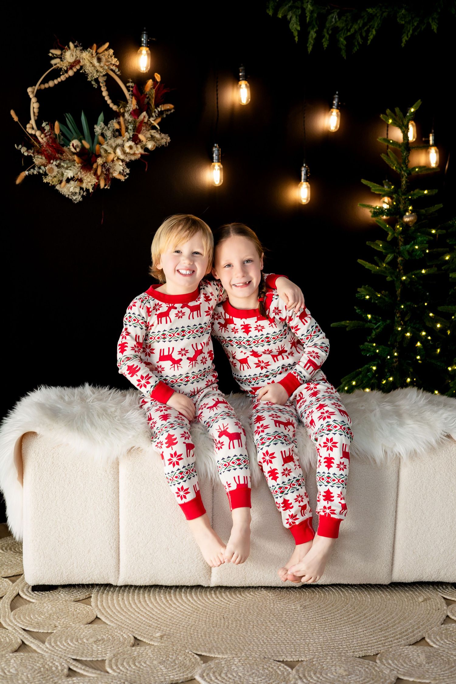 Two children in matching Christmas pajamas sit smiling on a white couch with holiday lights and tree.