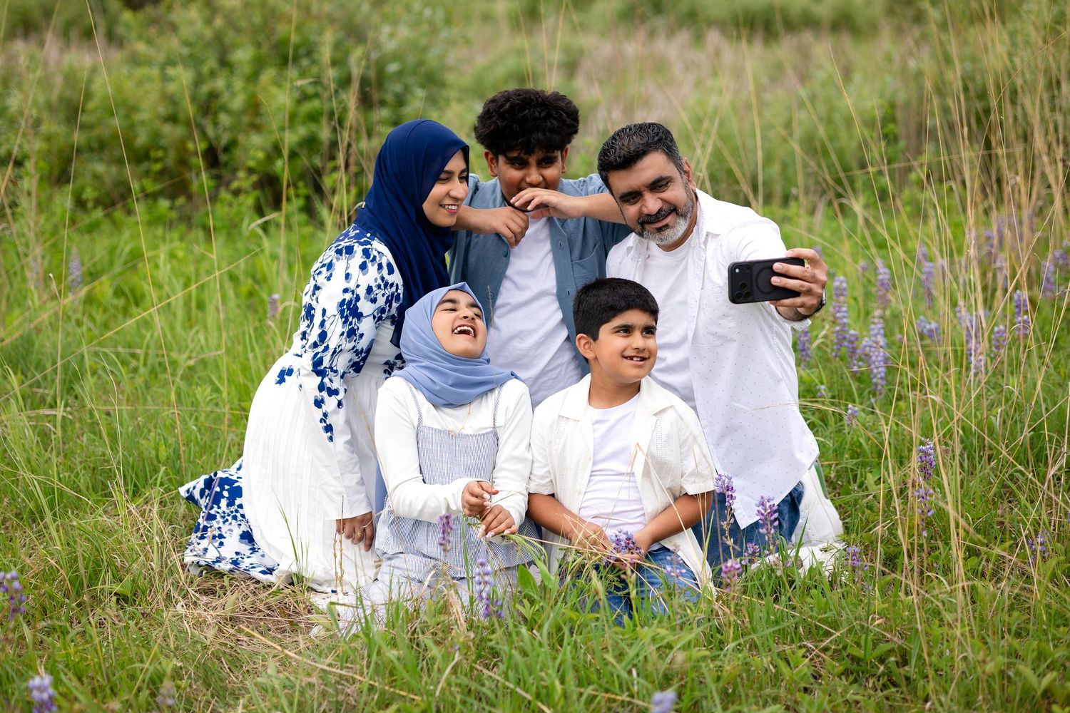 Muslim family taking a selfie together in a green field, smiling and laughing outdoors.