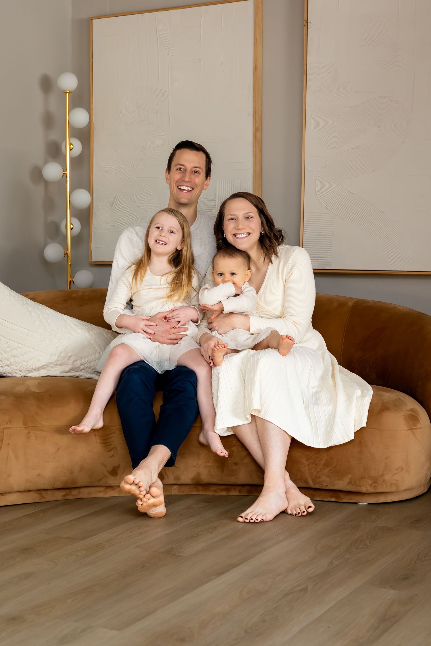 Happy family of four sitting on a brown velvet sofa, parents smiling with young daughter and baby in a bright room.