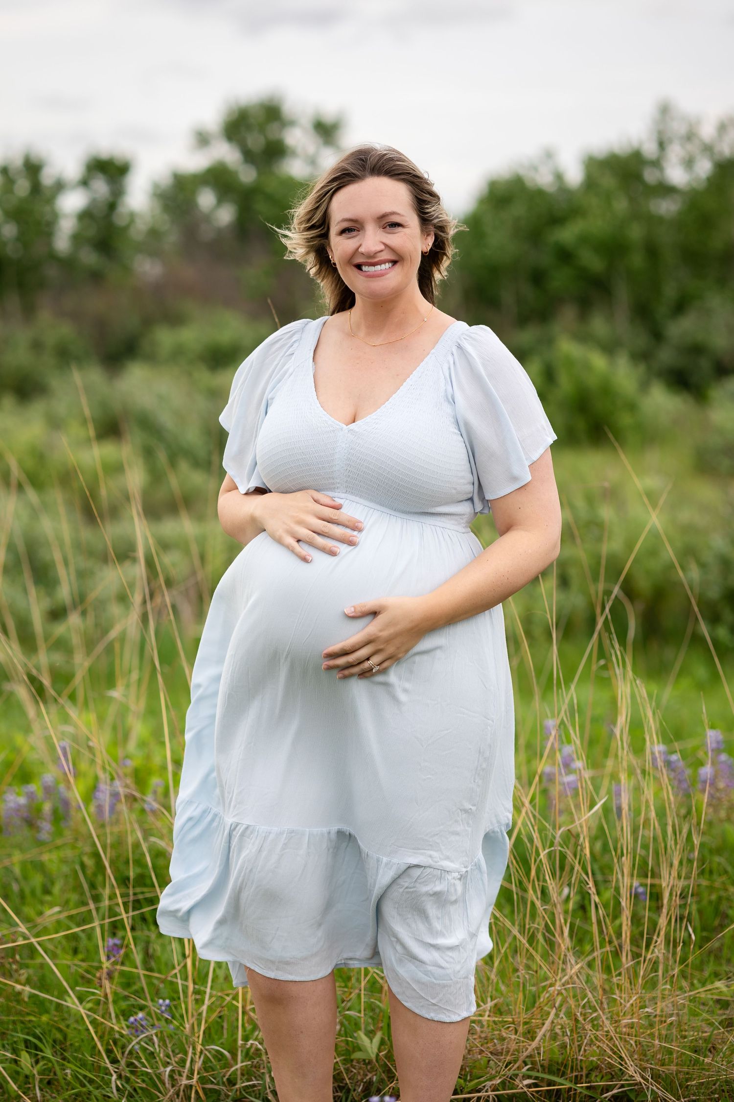 Smiling pregnant woman in white dress cradling baby bump outdoors in green field.