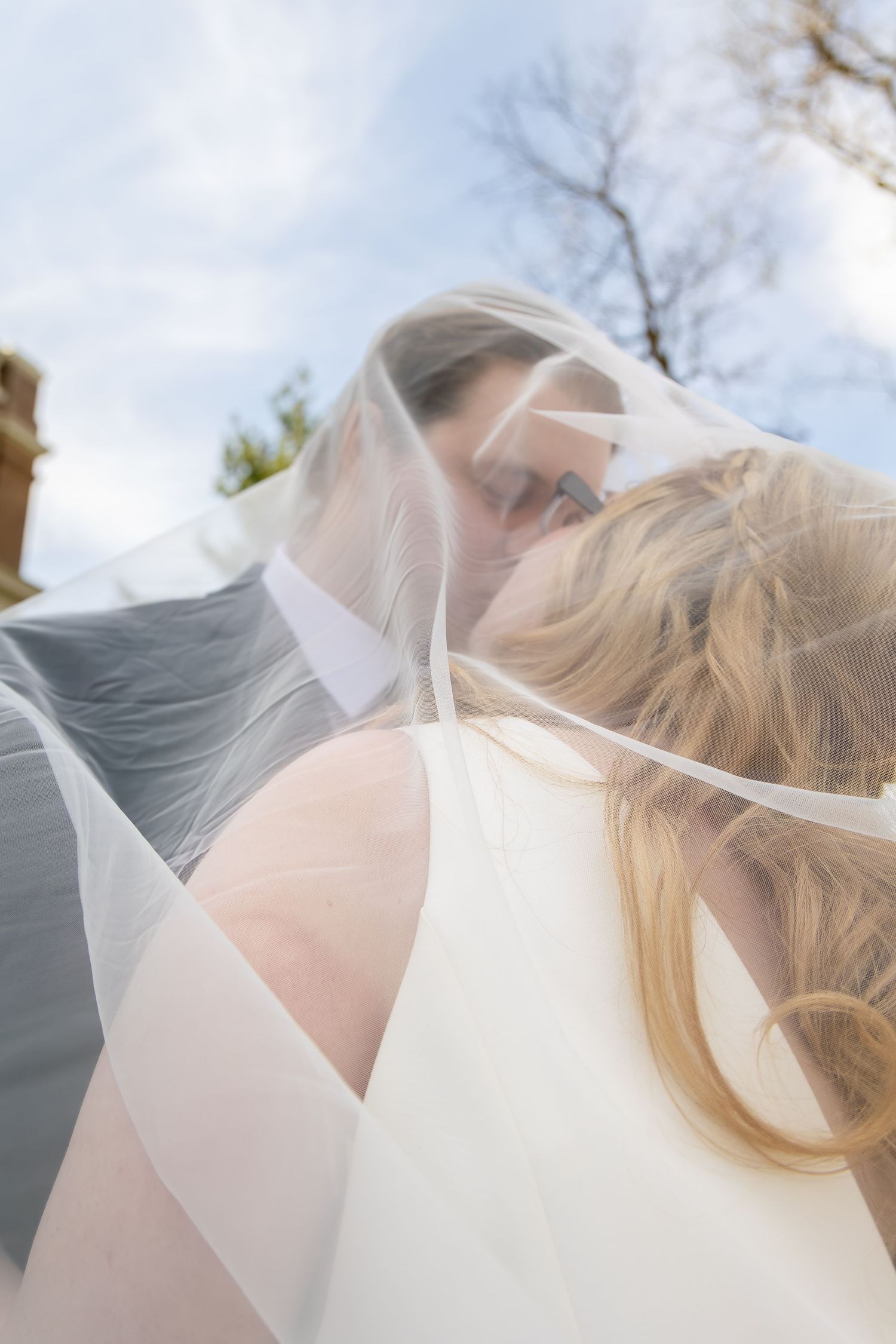 Romantic wedding couple sharing a kiss under a flowing bridal veil against a blue sky backdrop.