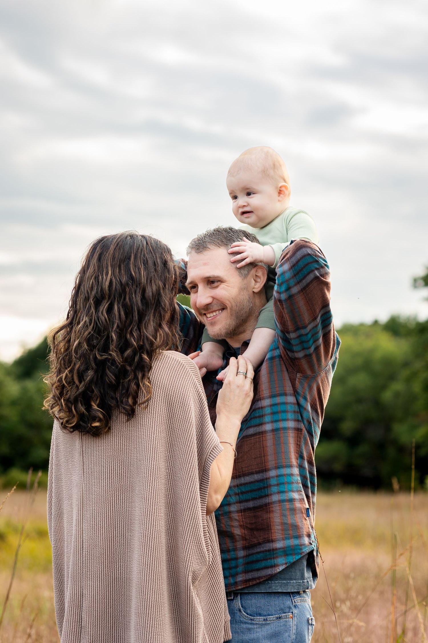 A family shares joyful moments together in a sunlit field with tall grass and cloudy skies.
