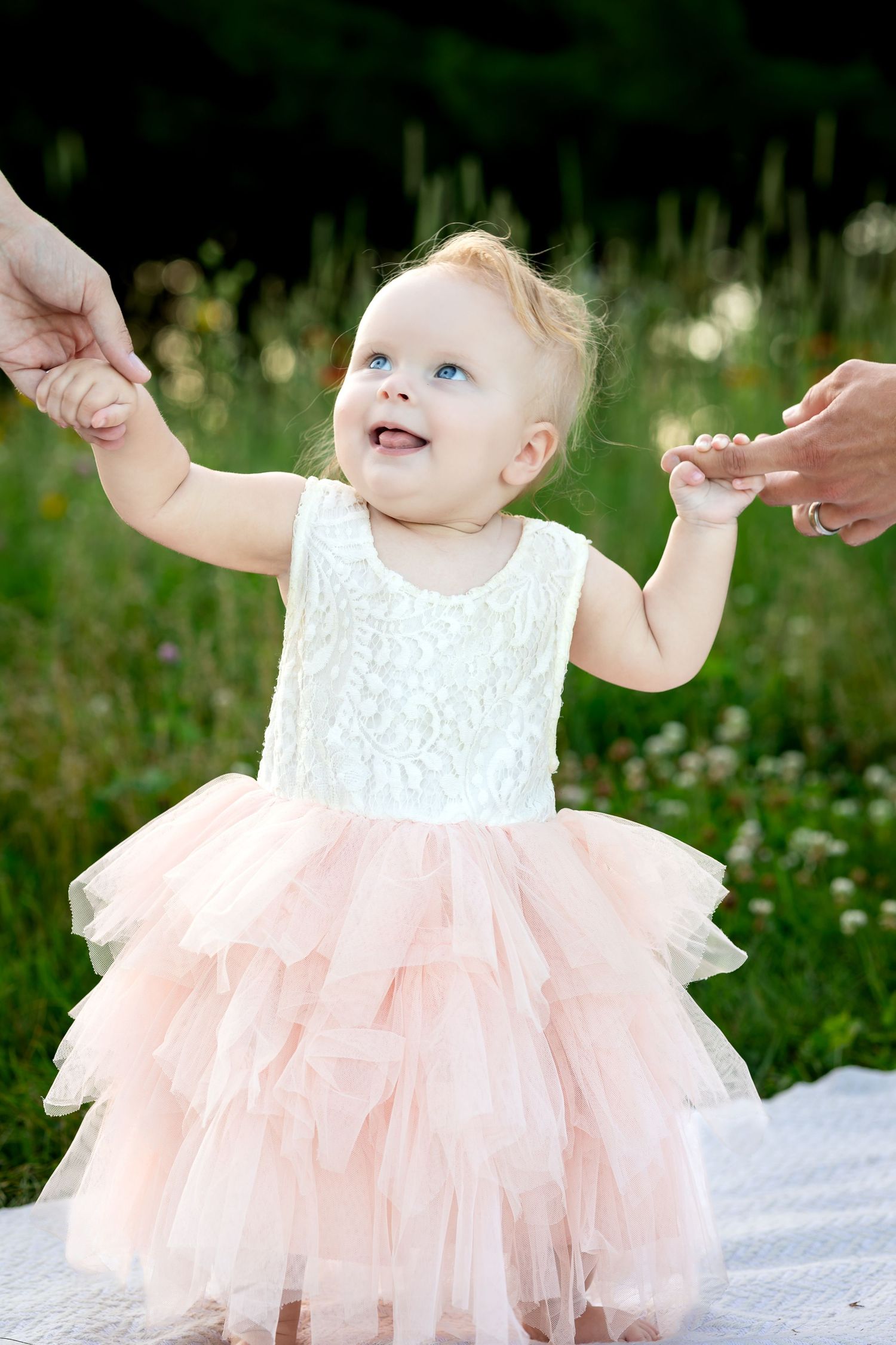 Small child in white and peach tulle dress stands in a meadow while holding adult hands on both sides.