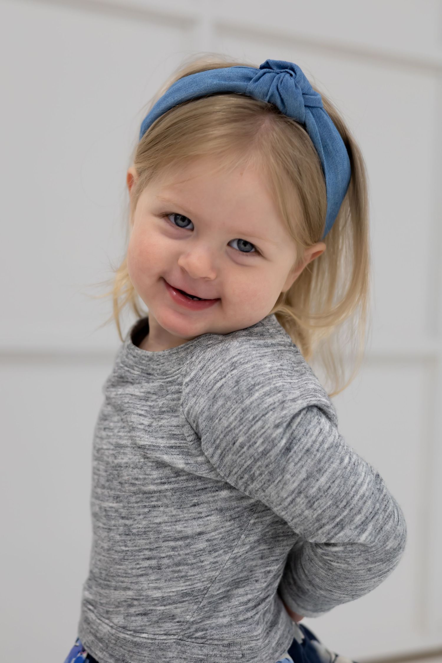 A small child wearing a blue headband and gray sweater smiles playfully against a white background.