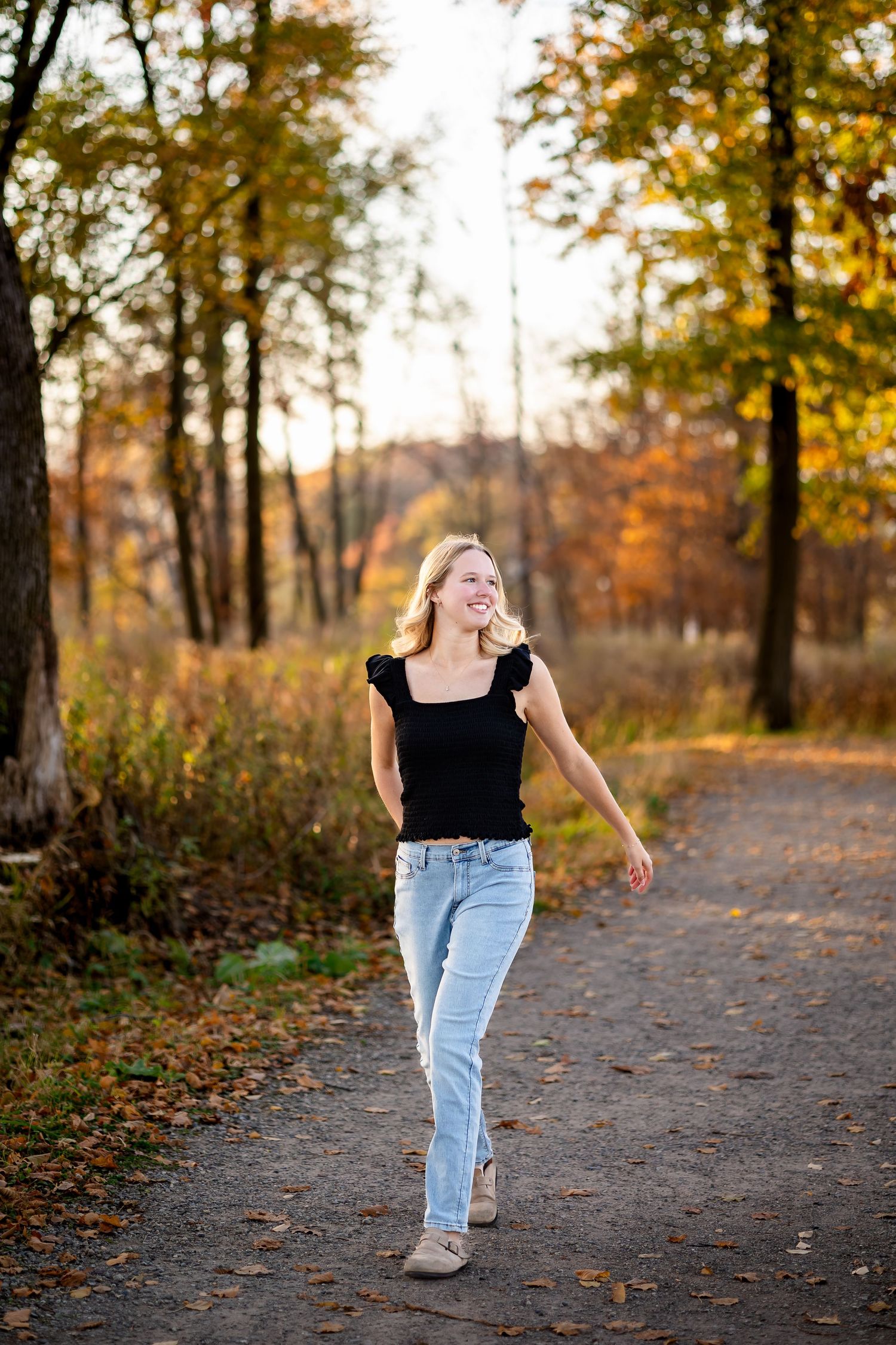 A person wearing jeans and a black top walks along an autumn forest path with fallen leaves at sunset.
