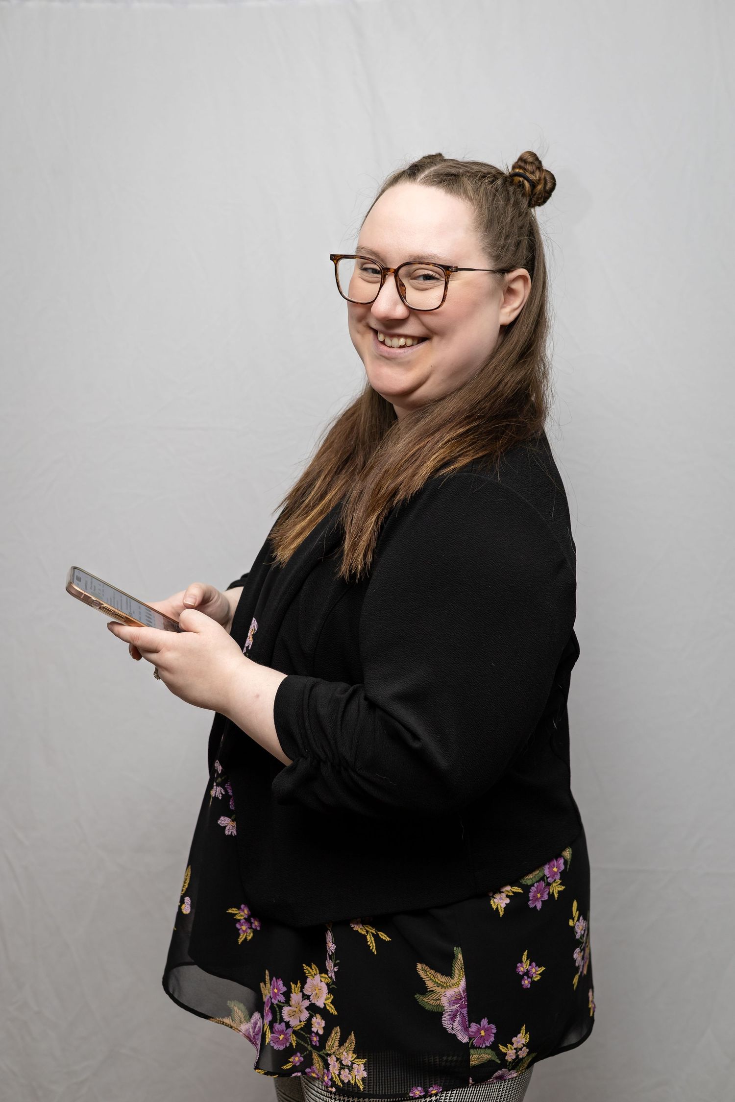 A person in glasses and a black floral dress smiles while holding a mobile phone against a gray background.