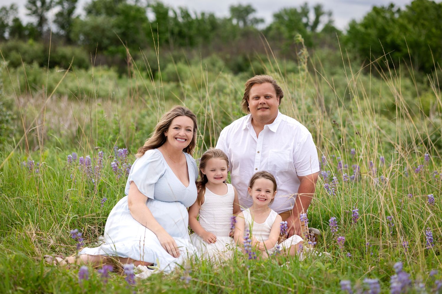 Family of four smiling together in a wildflower meadow, mother in blue dress, father in white shirt, two young daughters.