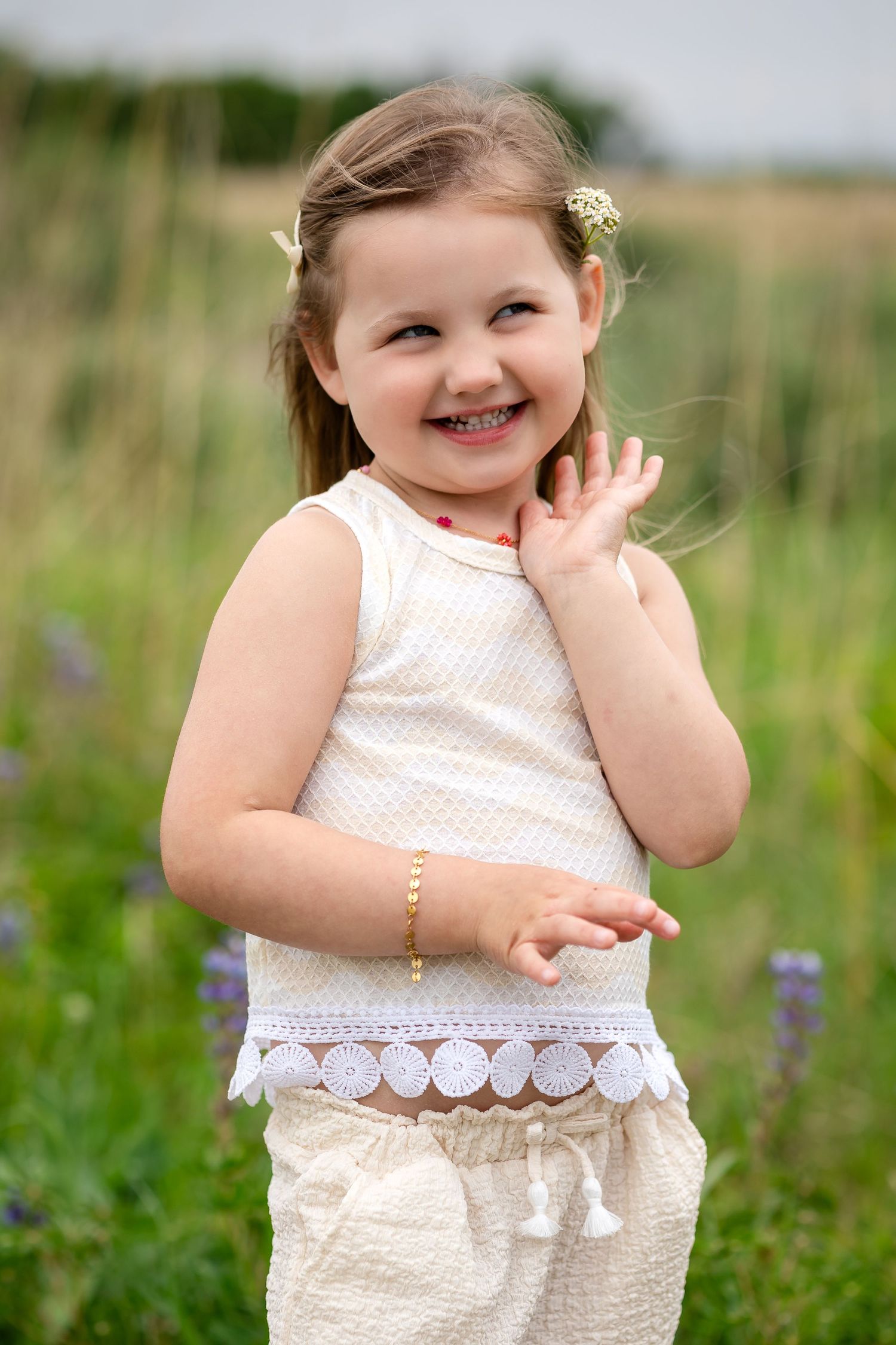A cheerful little girl in a white outfit smiles in a green field, wearing a bracelet and flower in her hair.