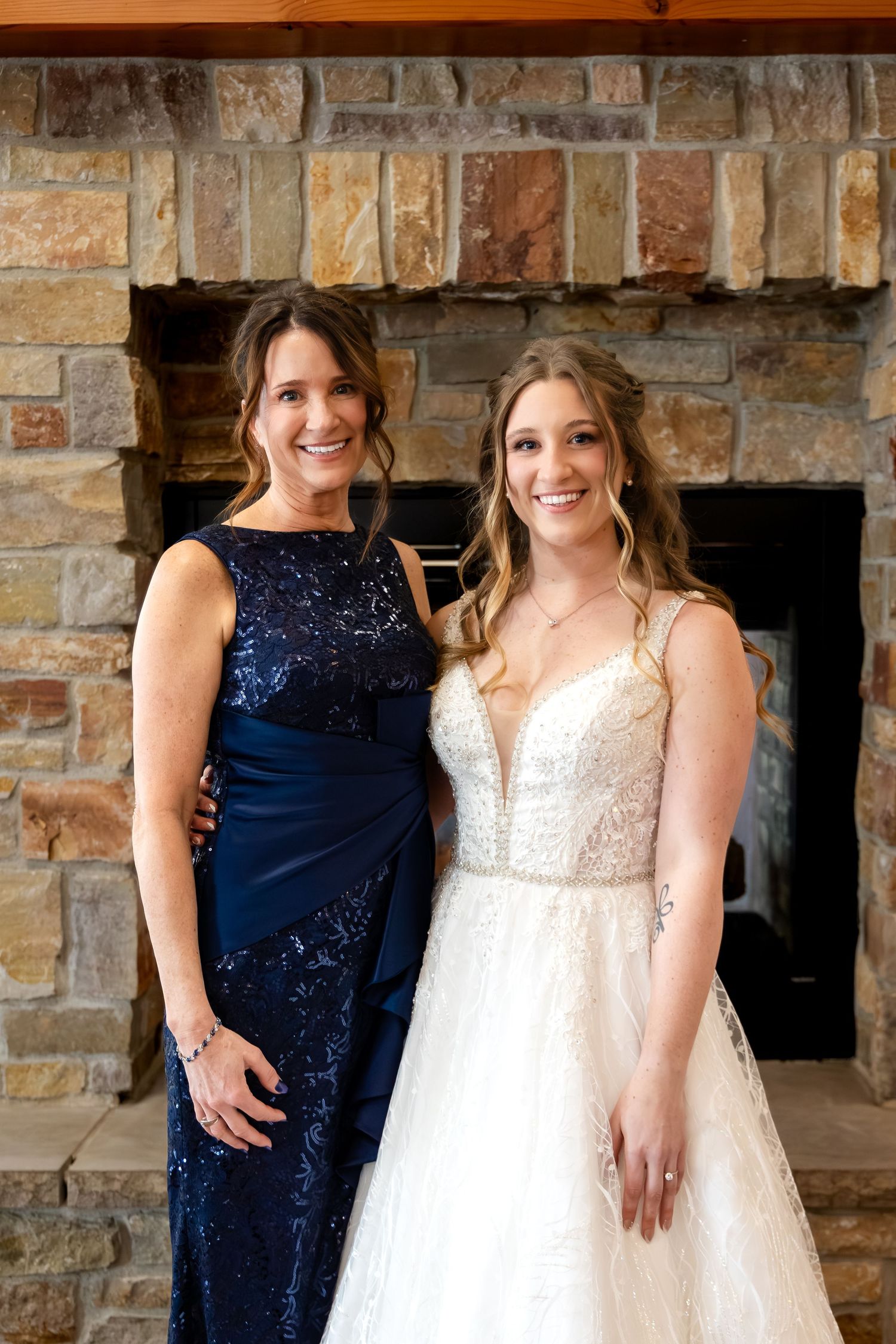 Bride in white lace gown poses with woman in navy sequin dress before stone fireplace.