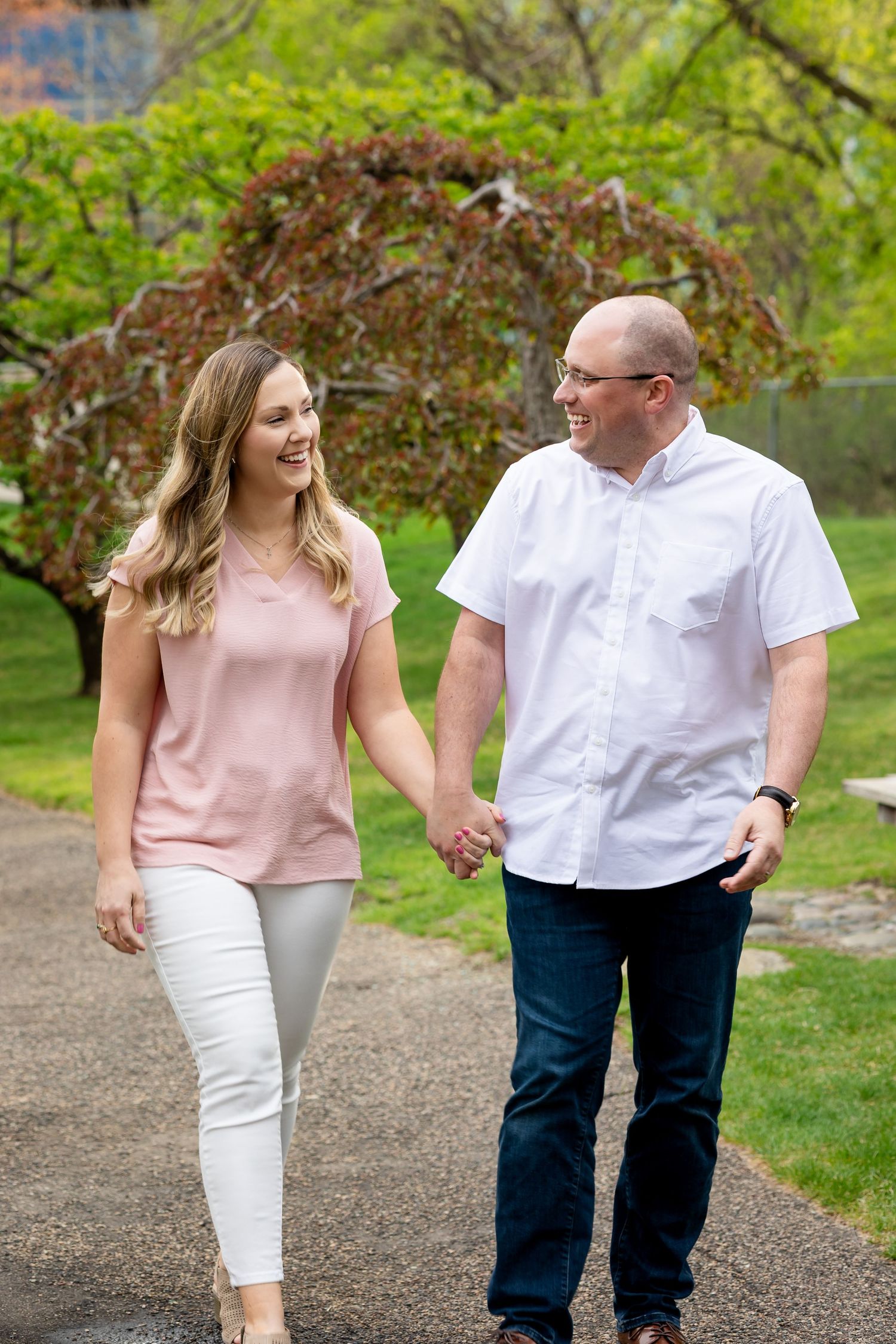 Smiling couple walking hand in hand on a path surrounded by vibrant green trees in a park.