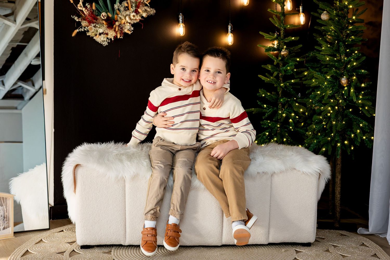 Two young boys in matching striped sweaters sit together on a white bench beside a decorated Christmas tree.