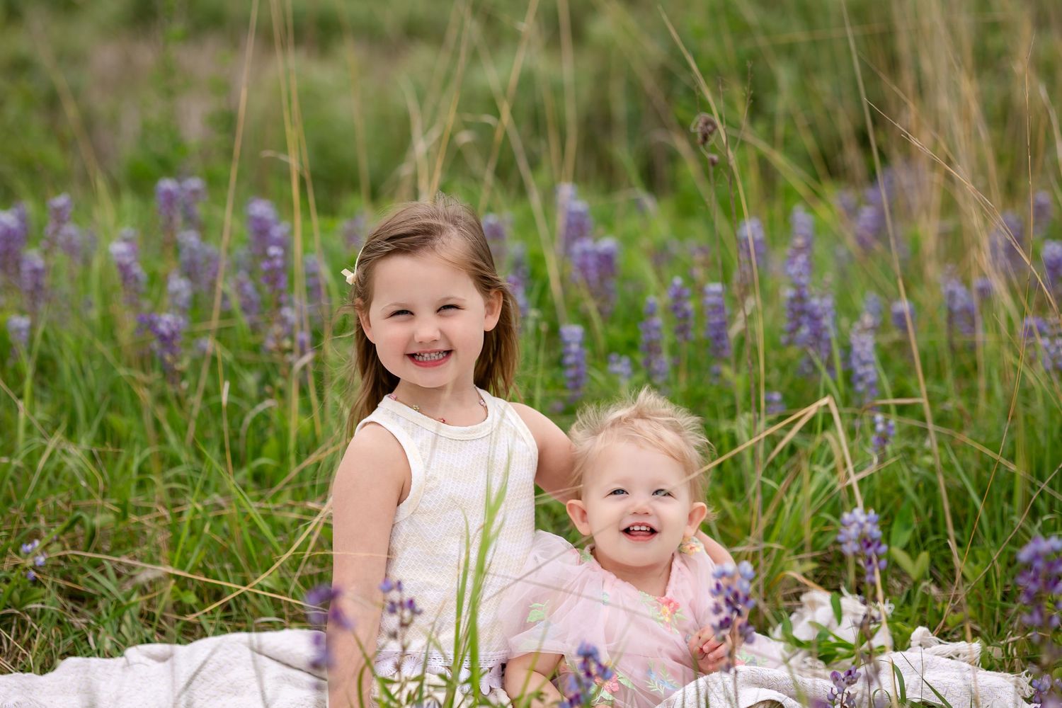 Two smiling young girls sitting in a field of purple wildflowers wearing white and pink dresses.